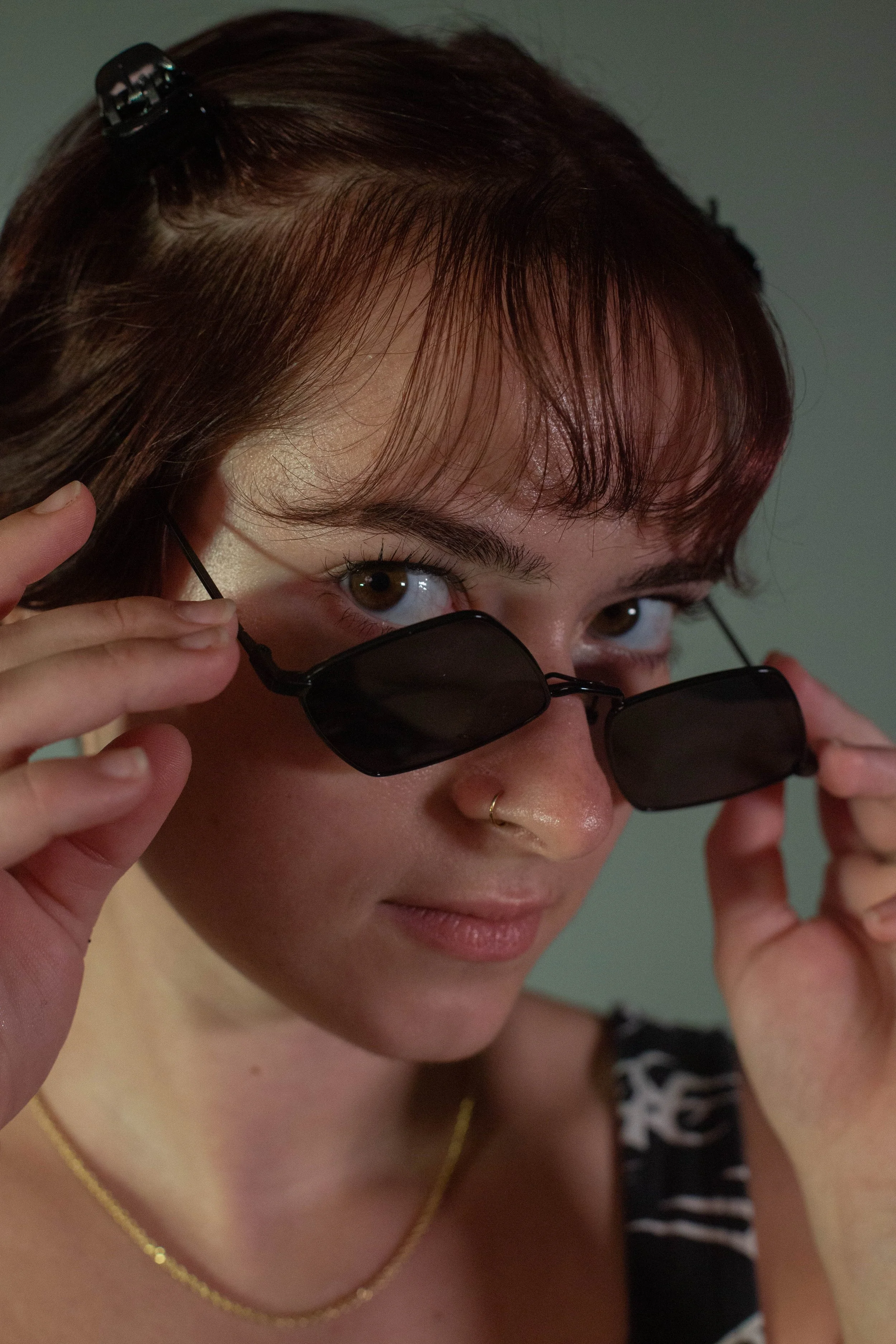 A young woman with brown hair and hazel eyes is pulling down black sunglasses to look directly at the camera.
