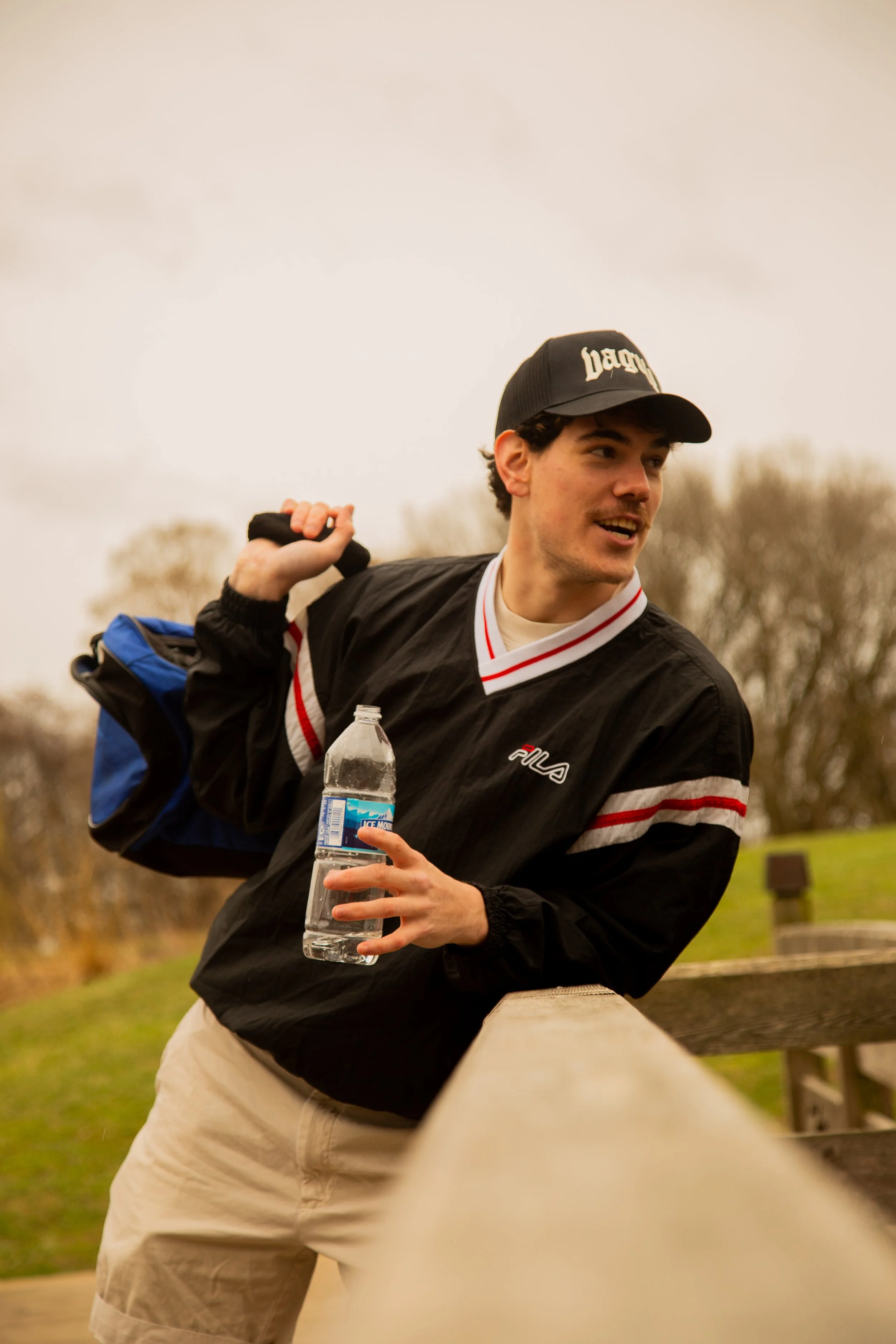 A young man with dark hair wearing a black and red FILA windbreaker, a black cap, and beige shorts, leaning on a wooden railing outdoors on a cloudy day. He is holding a water bottle and a blue and black backpack over his shoulder, smiling and lookin