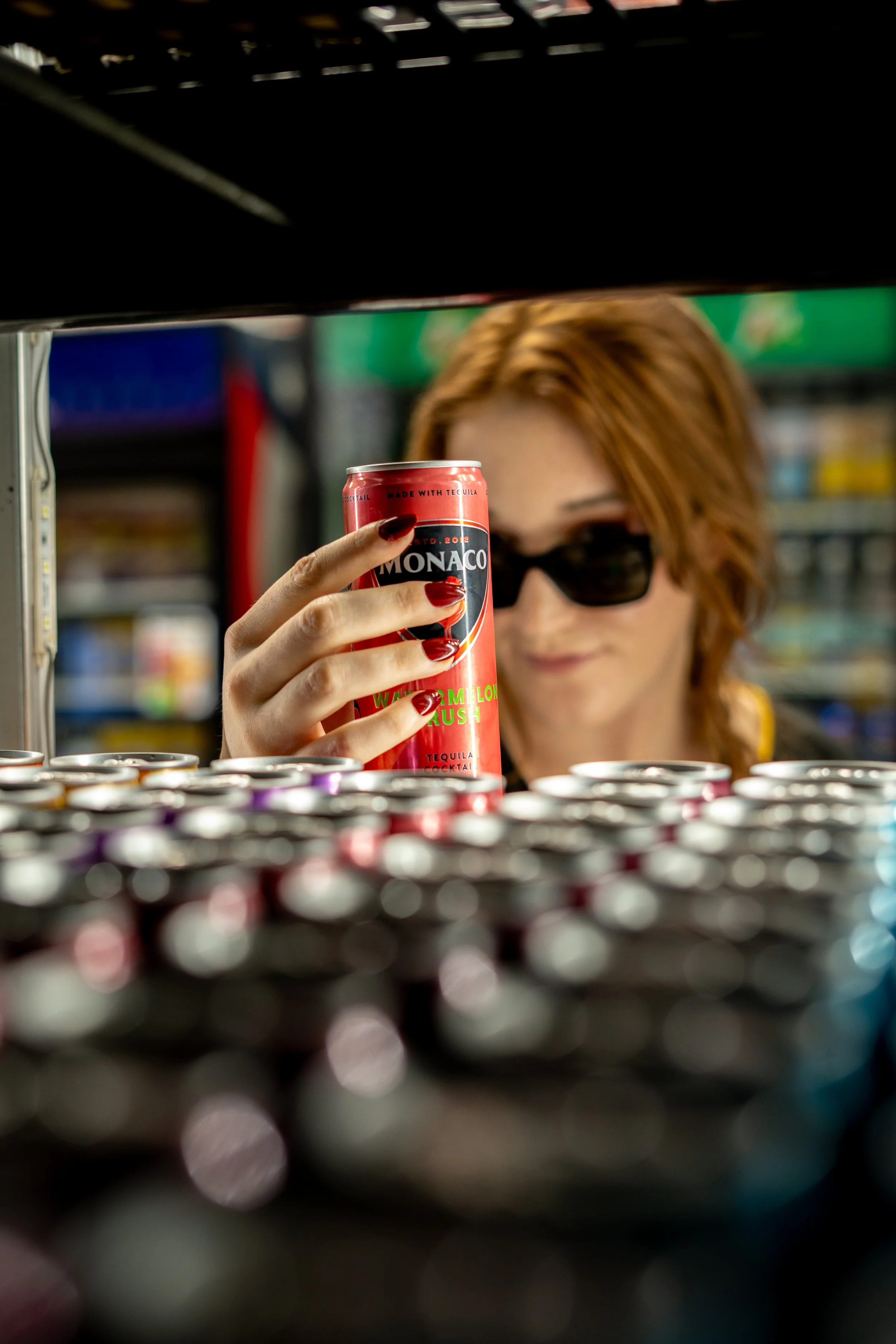 A woman with reddish hair and sunglasses holding a can of Monaco Waui Melon Rush drink while in a store aisle, viewed from below through a display of canned beverages.