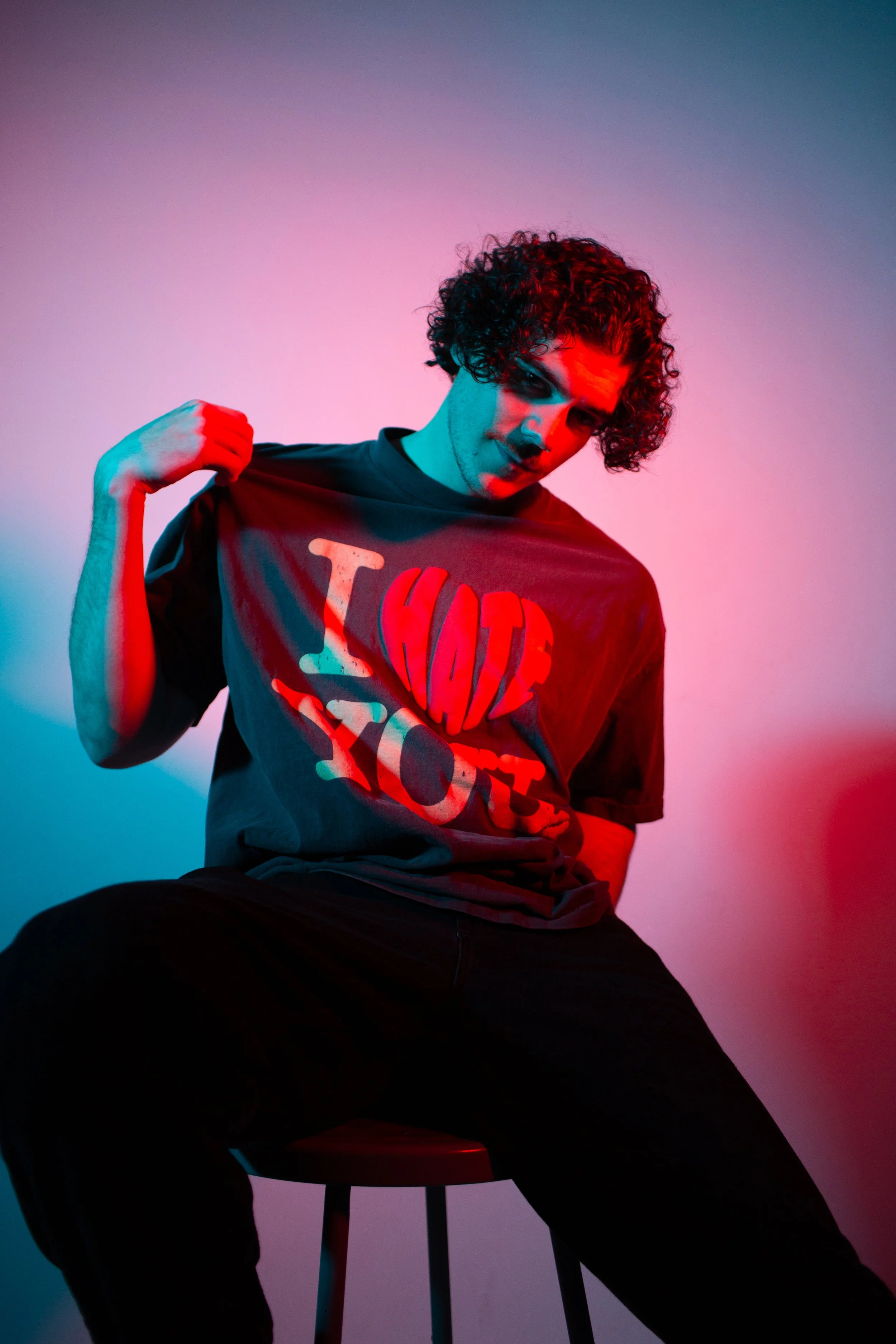 A young man with curly hair sitting on a stool in colorful lighting, holding up his T-shirt to display a graphic design.