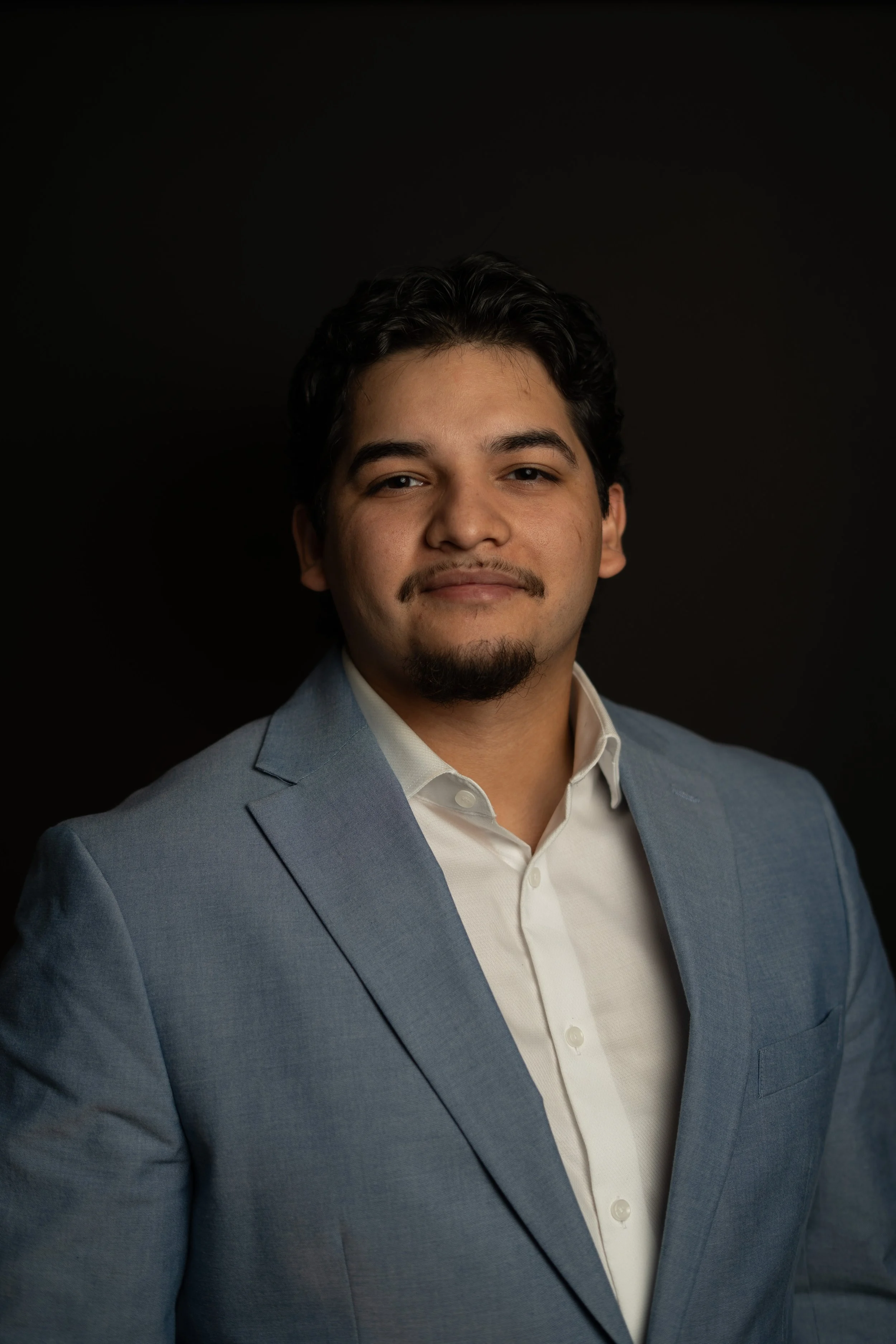 Headshot of a young man with dark hair, a mustache and goatee, wearing a light blue blazer over a white shirt, against a black background.