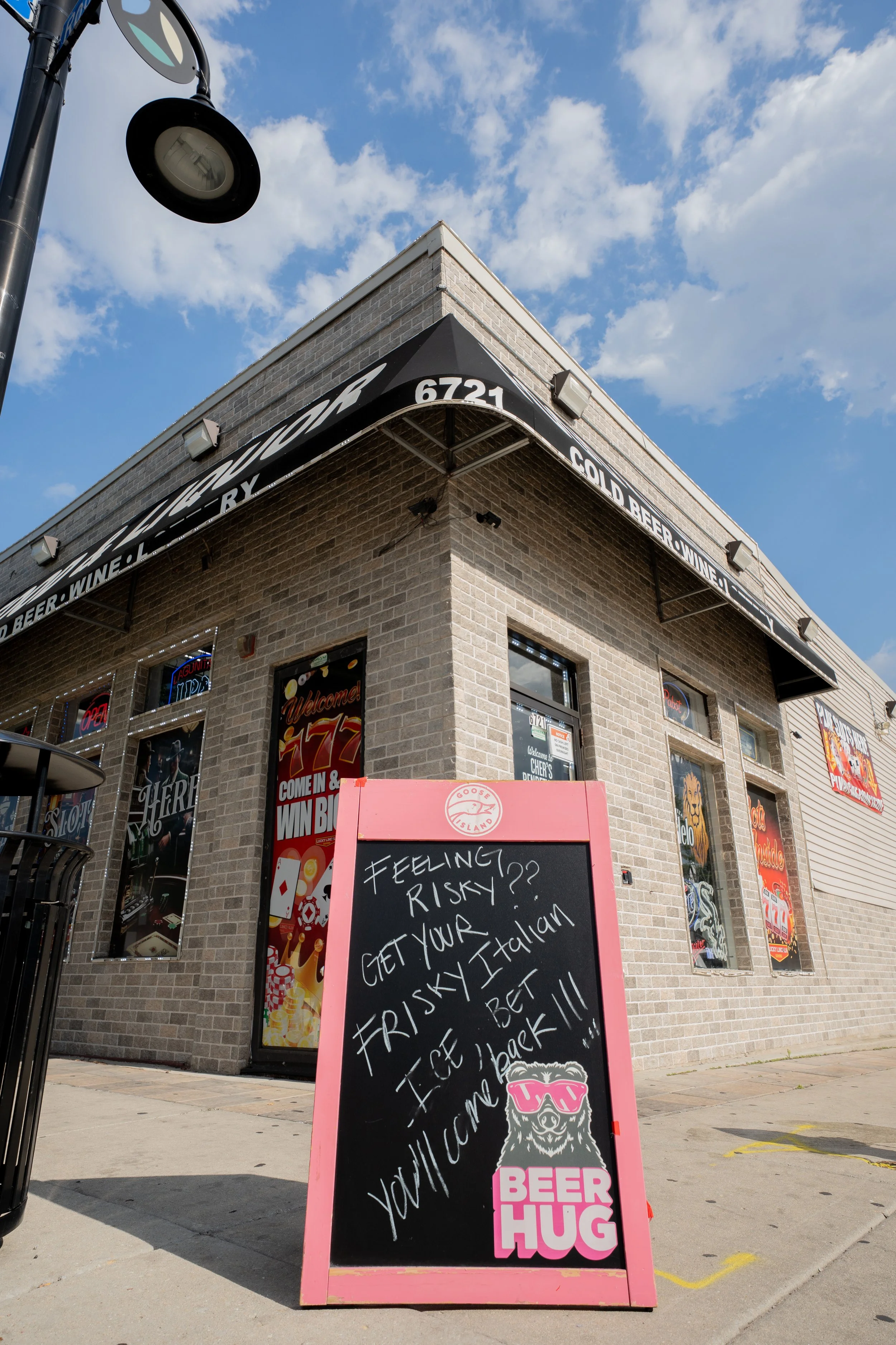 A sidewalk outside a brick building with large windows advertising beer and gambling; a pink sign with a cartoon bear wearing sunglasses and the words "BEER HUG" displays a handwritten message promoting a risky Italian beer, encouraging people to com