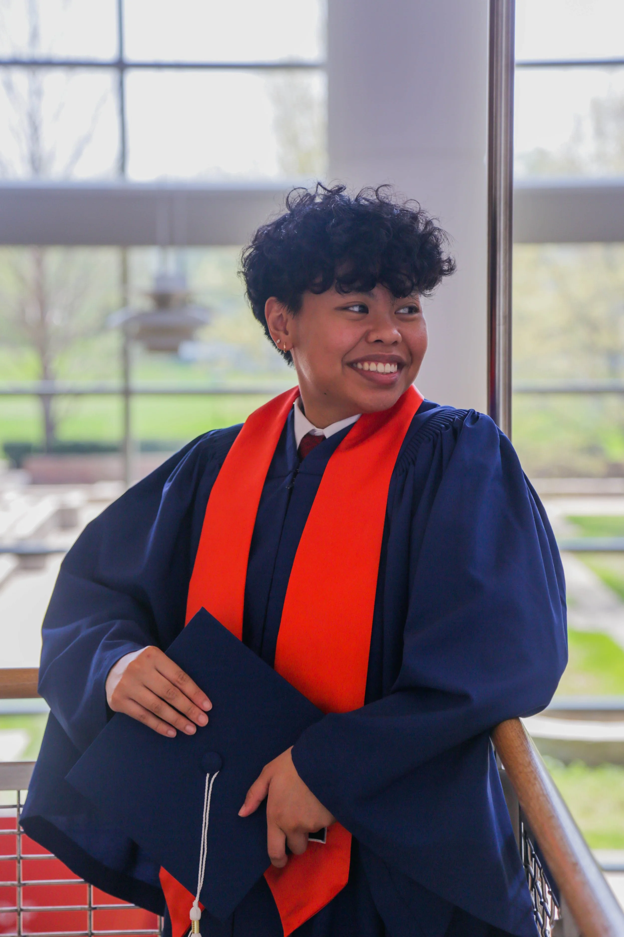A young woman at her graduation ceremony, wearing a dark blue cap and gown with an orange stole, holding her mortarboard, smiling and looking to her right in a room with large windows and a view of trees outside.