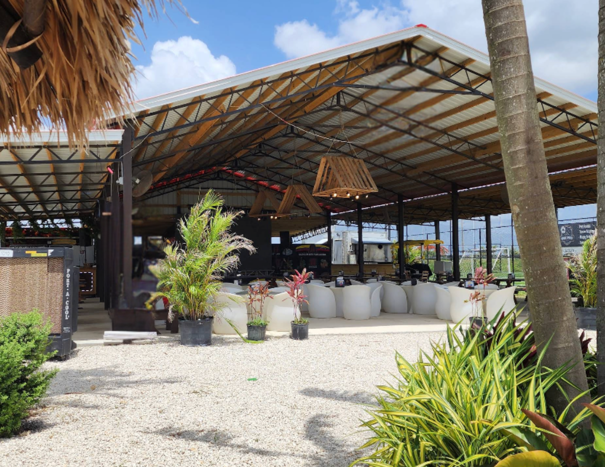 Outdoor seating area with white chairs and potted plants under a covered structure with a wooden and metal roof, surrounded by palm trees and greenery.