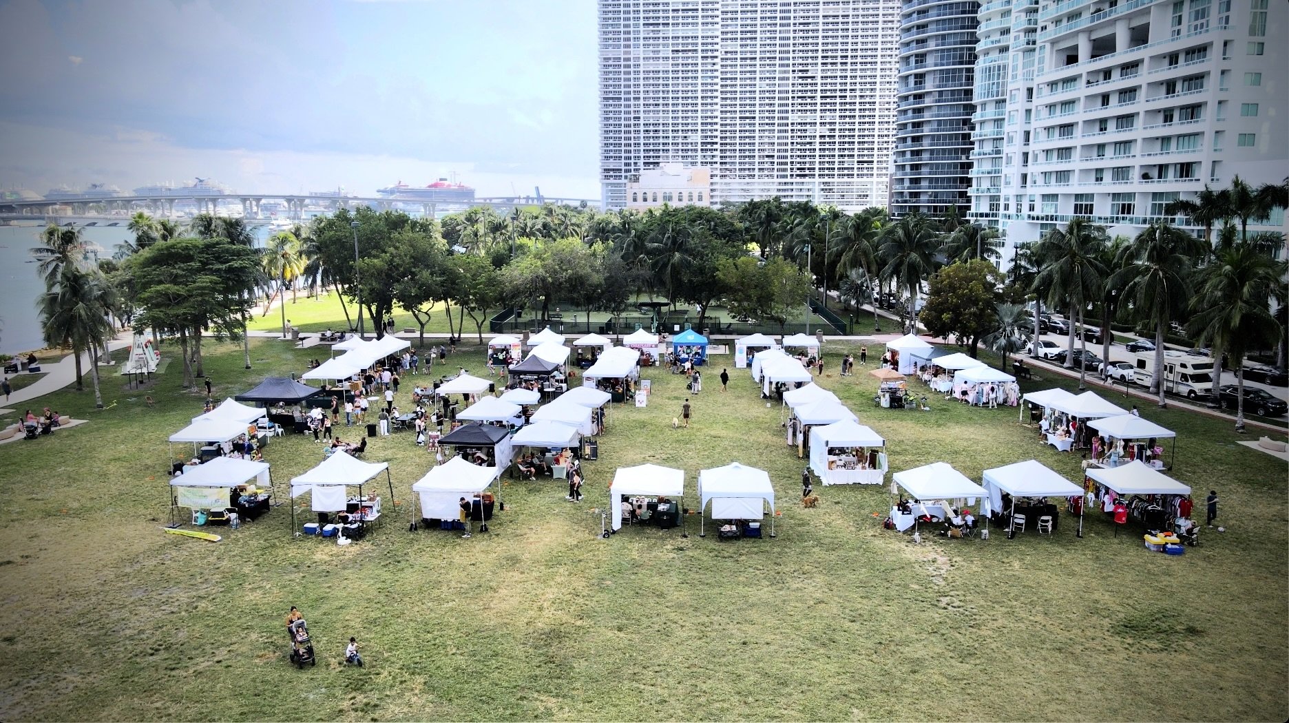 Outdoor market with white tents set up on a grassy area in front of a park with trees, palm trees, and high-rise buildings, with ships in a harbor in the background.