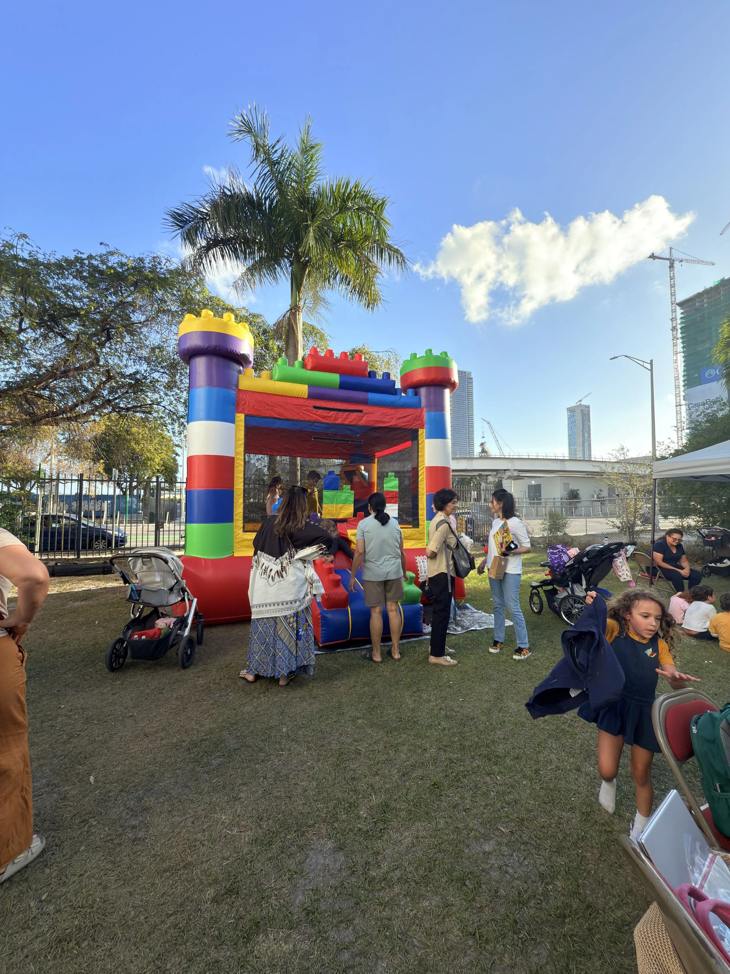 Children and adults gathered around a colorful inflatable bounce house at a park on a sunny day, with a palm tree and city buildings in the background.