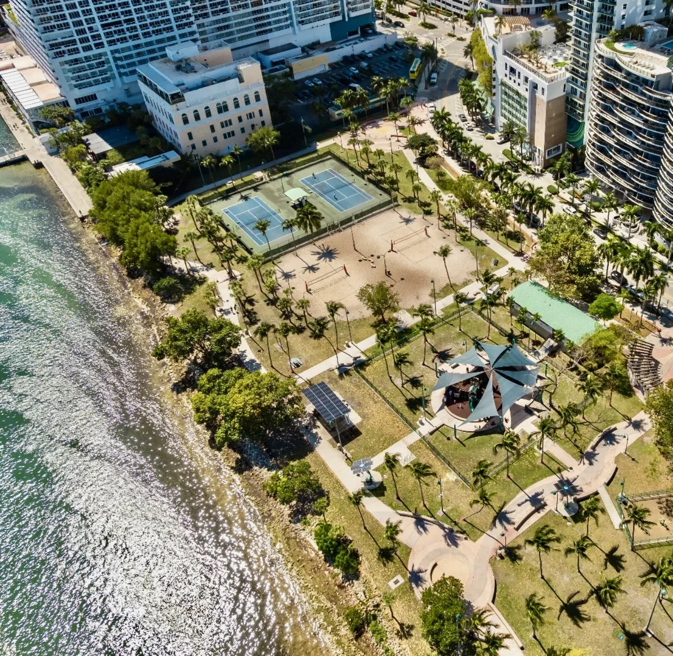 An aerial view of a waterfront park with tennis courts, a sandy volleyball court, walking paths, a shaded pavilion, and surrounding high-rise buildings.