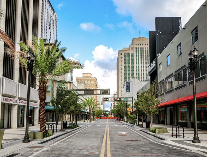 Empty city street with palm trees, tall buildings, and a blue sky with clouds.