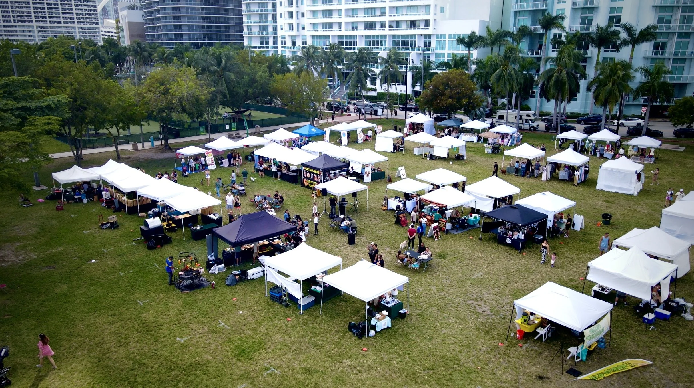 An outdoor market event with multiple white and a few black tents set up on a grassy area in front of modern high-rise buildings, with people walking and browsing various stalls.