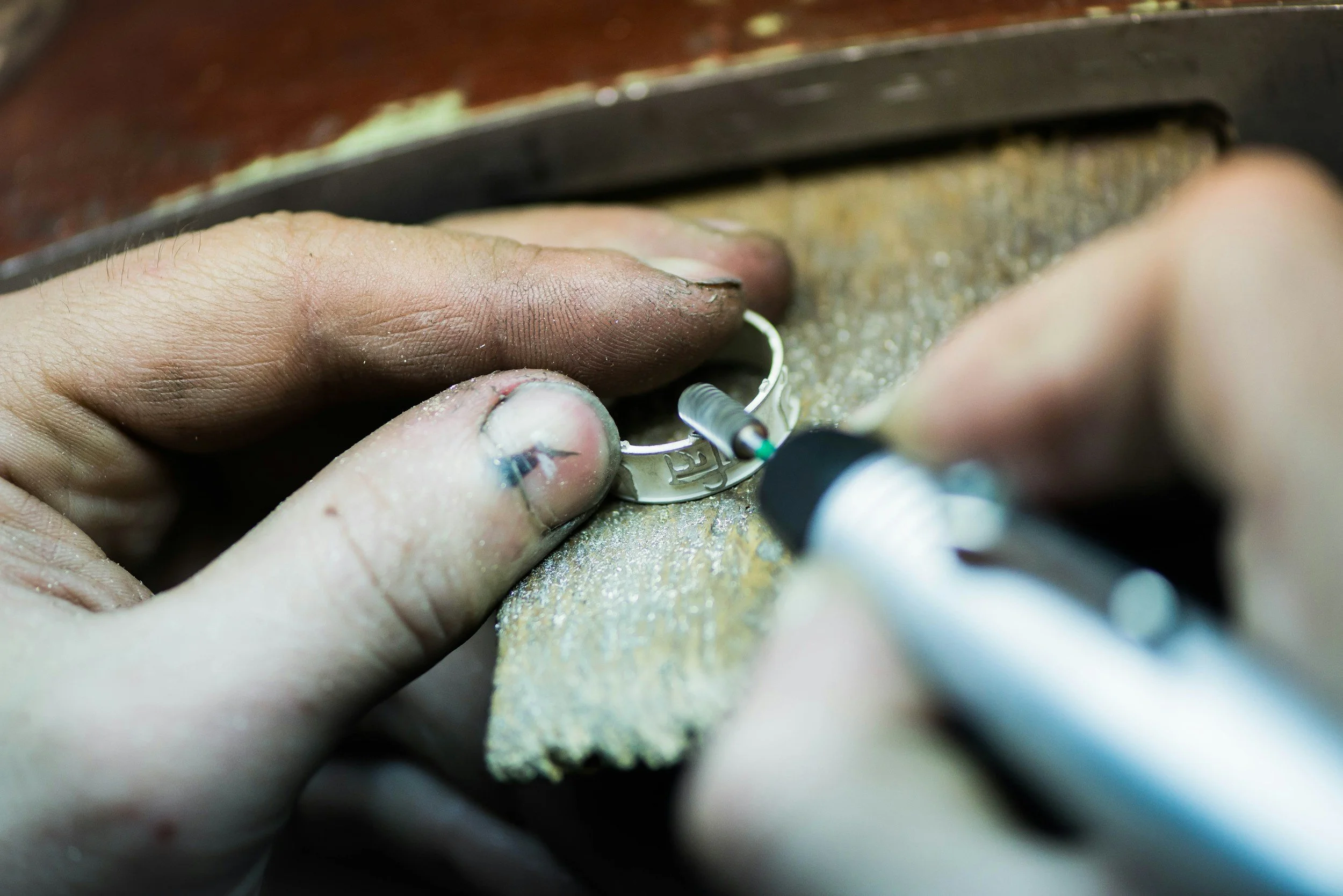 Close-up of hands carving or engraving a ring with a rotary tool. The fingers are dirty and the work surface is scattered with dust or debris.