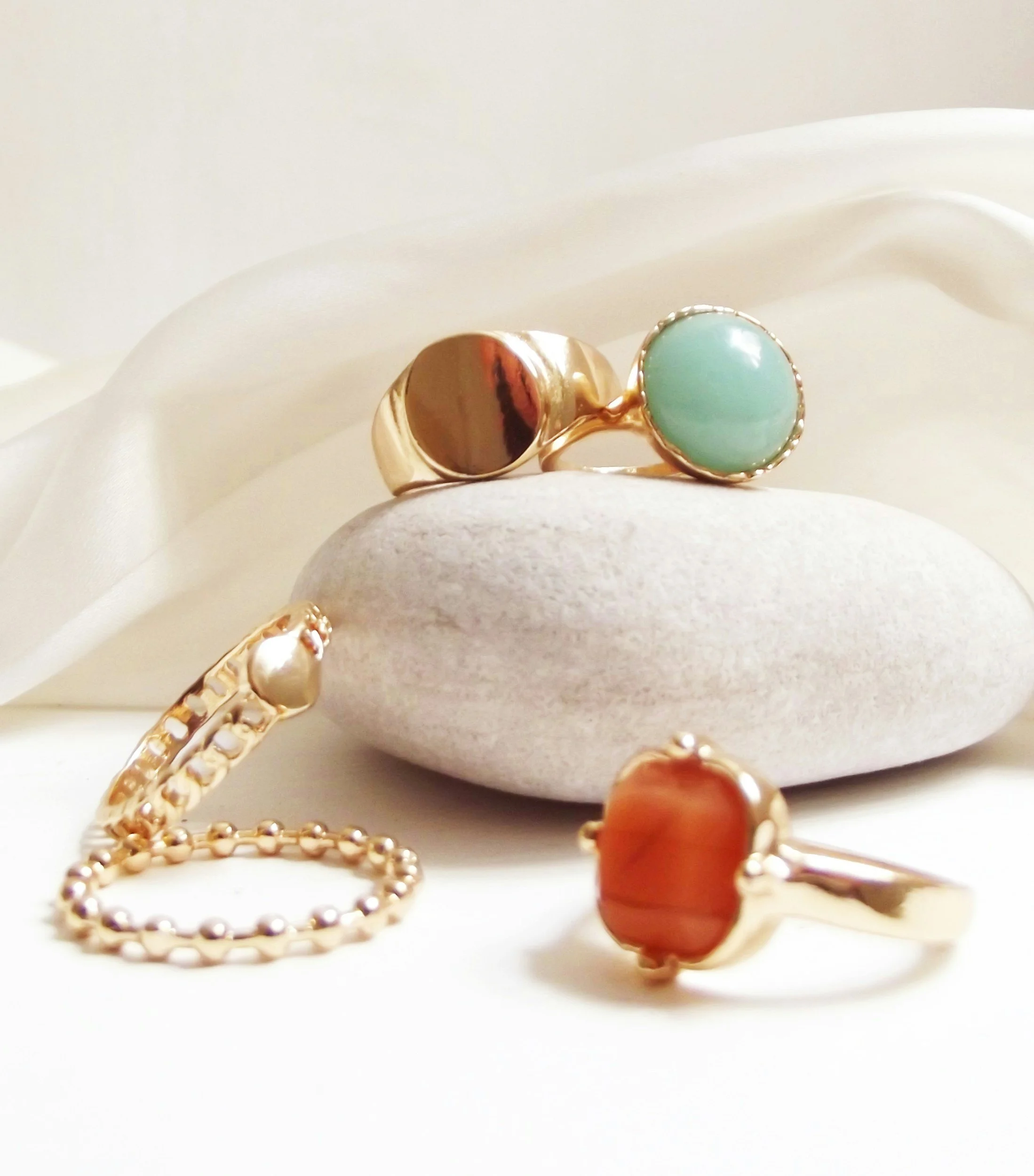 Close-up of gold jewelry including rings and a bracelet with colorful stones, placed on smooth white stones and draped white fabric.
