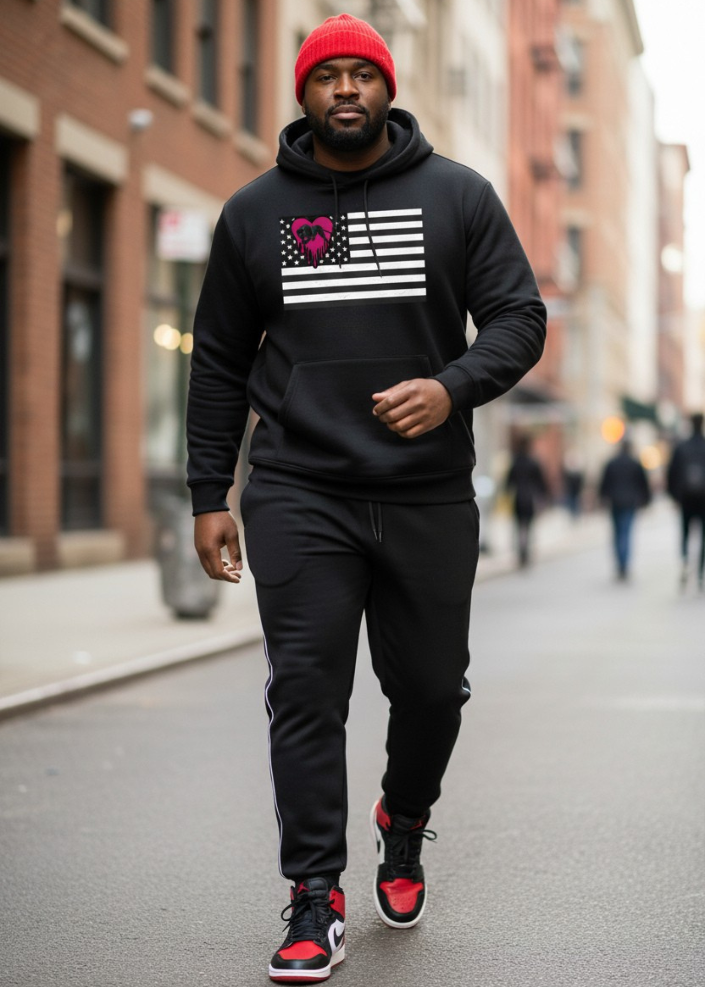 A man walking on city street wearing a black hoodie with a graphic of an American flag and a pink heart with glasses, black sweatpants, and red and black sneakers, with a red beanie and a beard.
