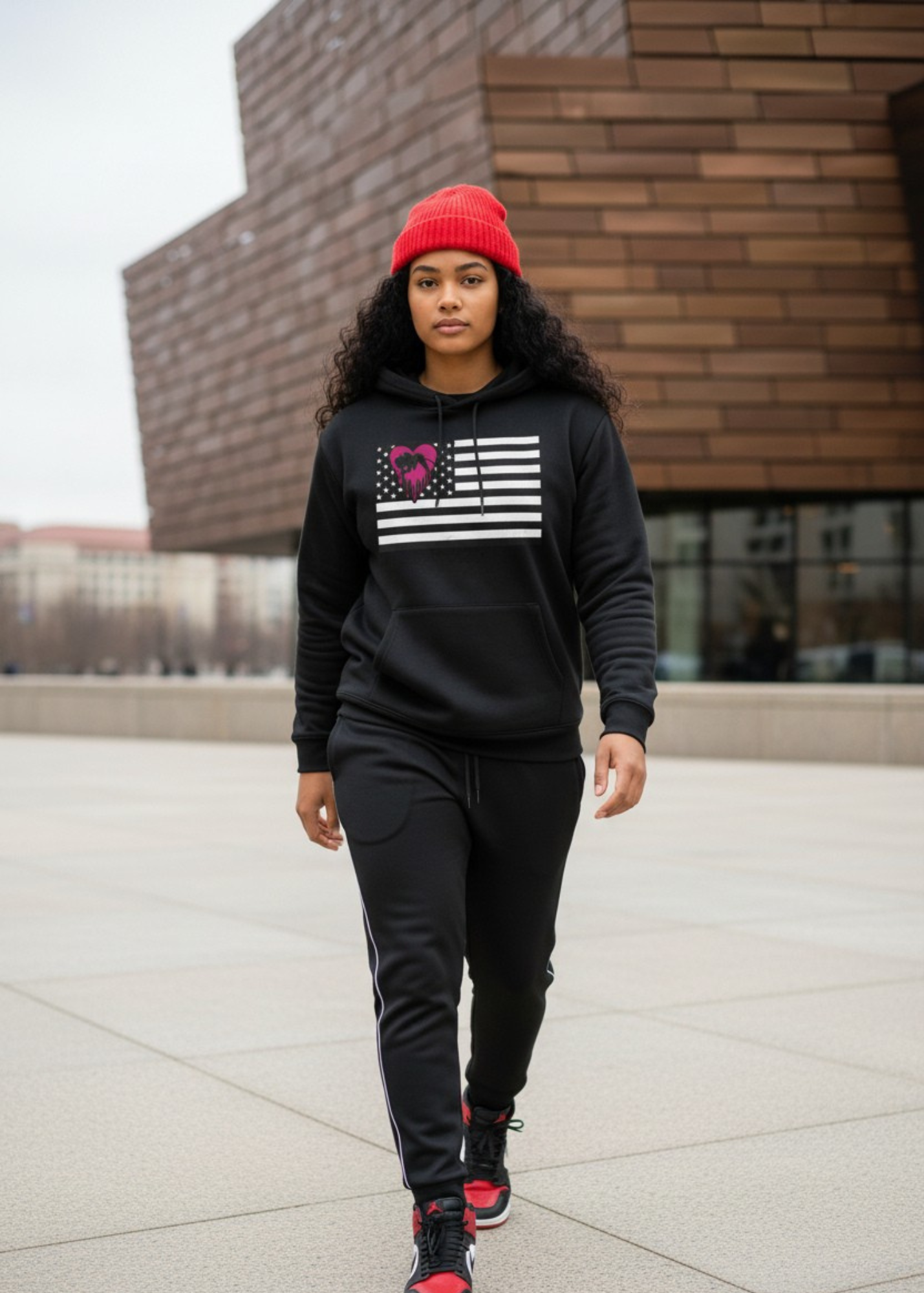 Young woman walking outdoors wearing a red beanie, black hoodie with a graphic of the American flag and a pink heart, black sweatpants, and black and red sneakers.