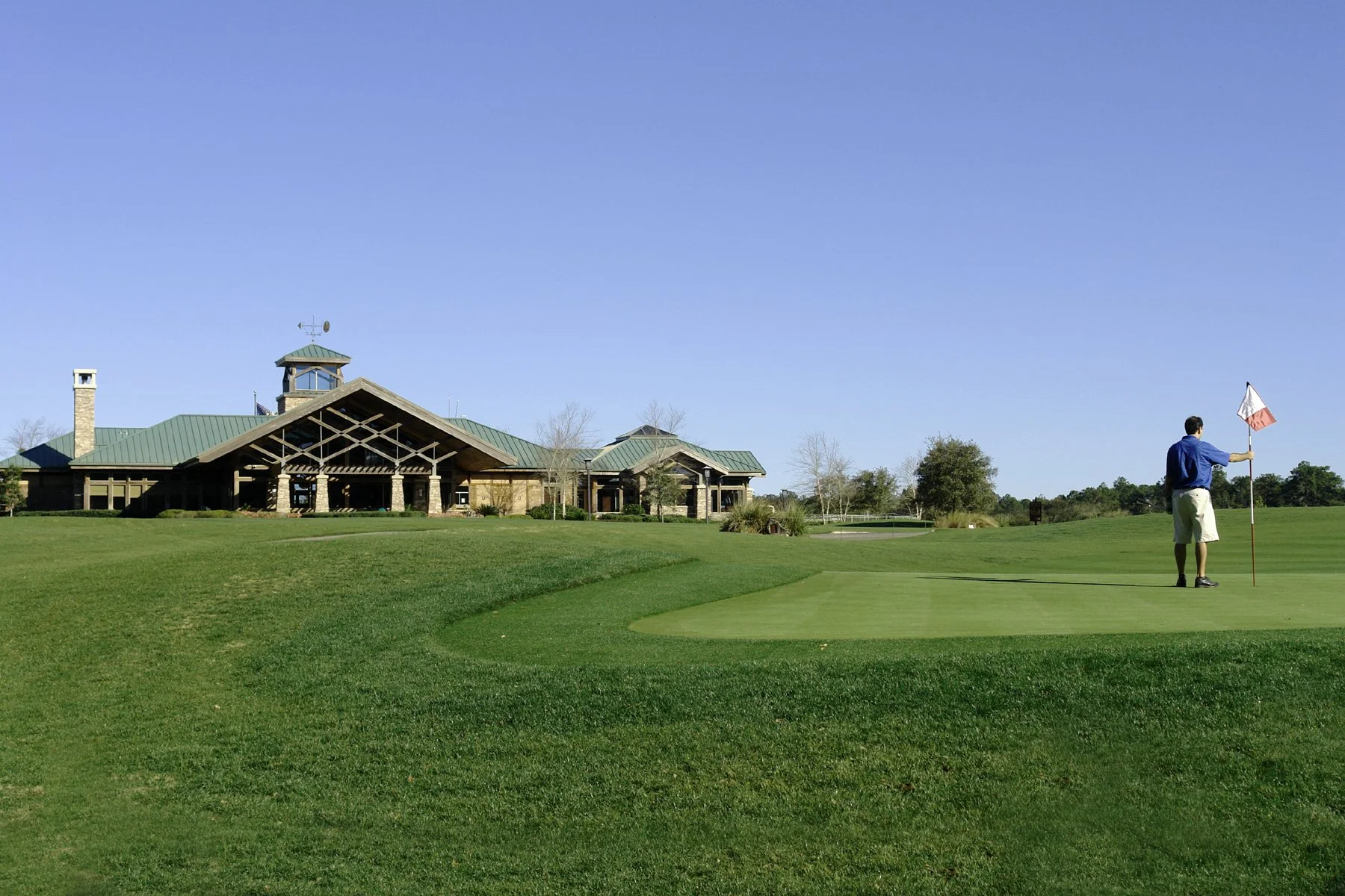 A man holding a golf flag on a putting green at a golf course with a clubhouse in the background under a clear blue sky.