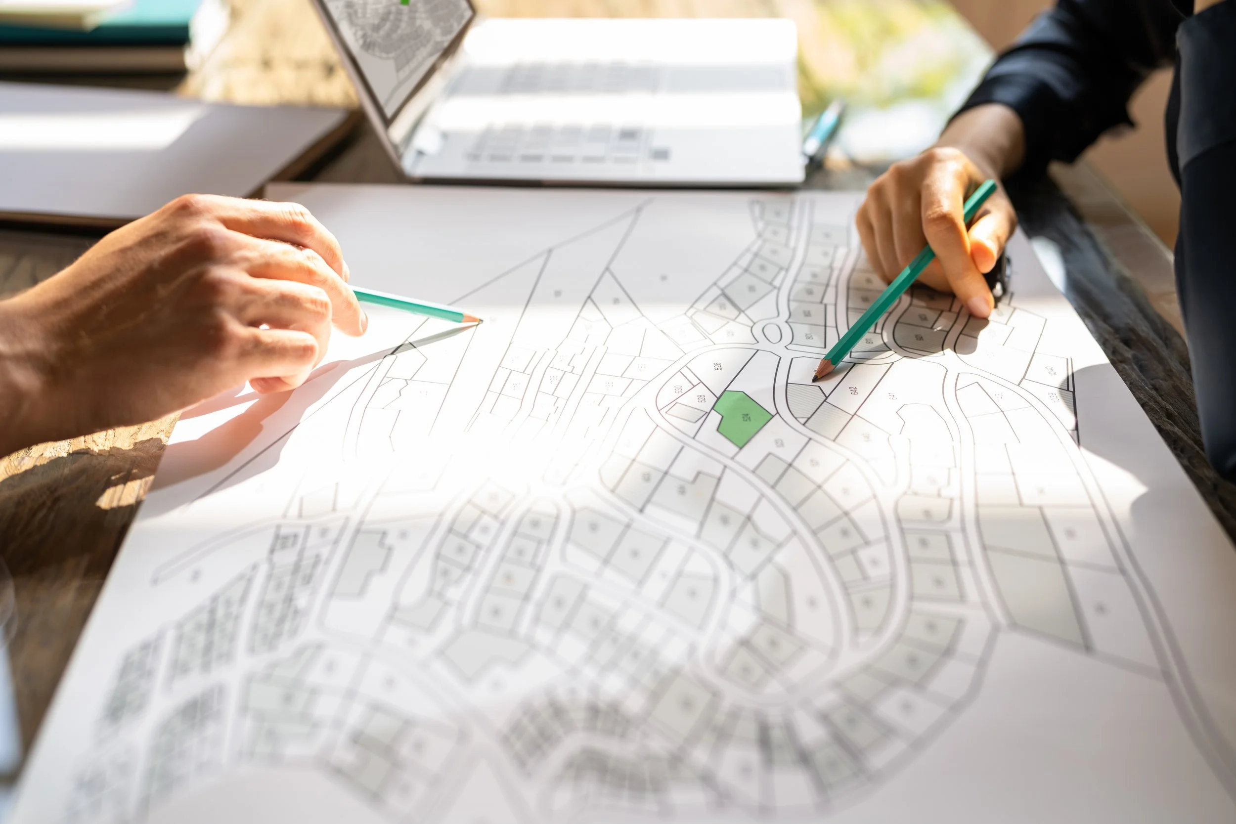 Two individuals analyzing a detailed neighborhood map with shaded areas, on a wooden table, with a laptop nearby.