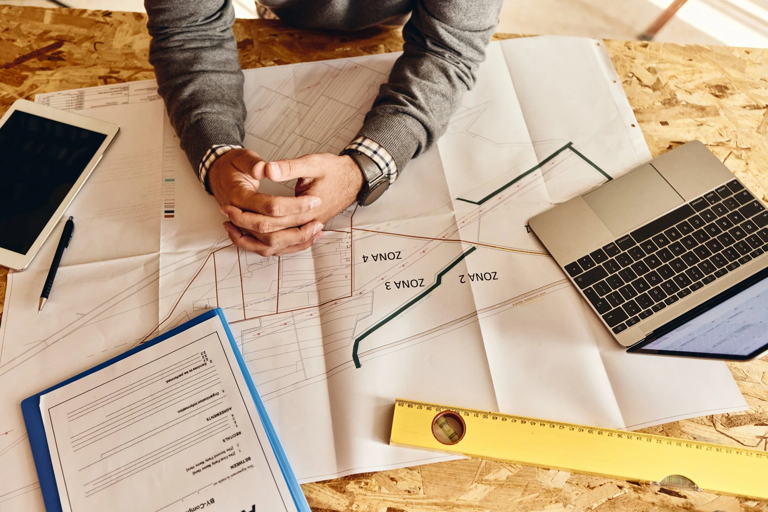 Person with folded hands sitting at a work table with blueprints, a laptop, a tablet, a pen, a ruler, and a clipboard.