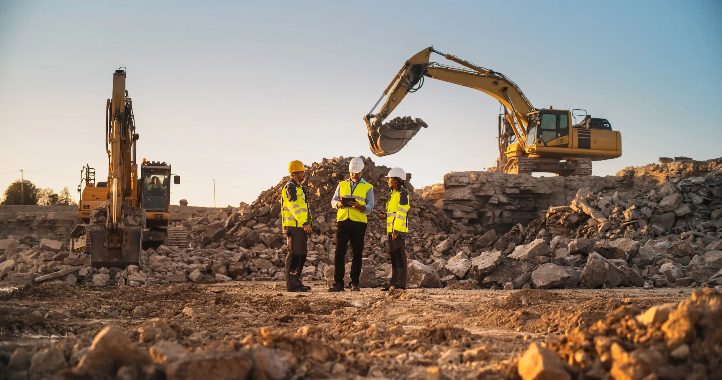 Construction site with two excavators moving rocks and three construction workers in safety vests and helmets discussing plans, during sunset.