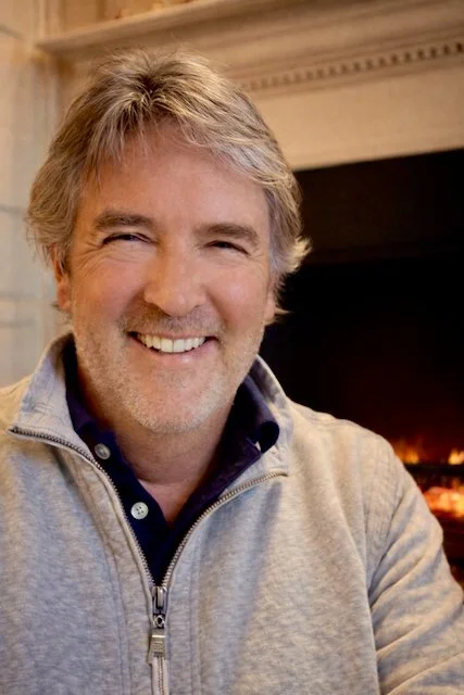 A middle-aged man with gray hair and a beard smiling, sitting indoors in front of a fireplace.