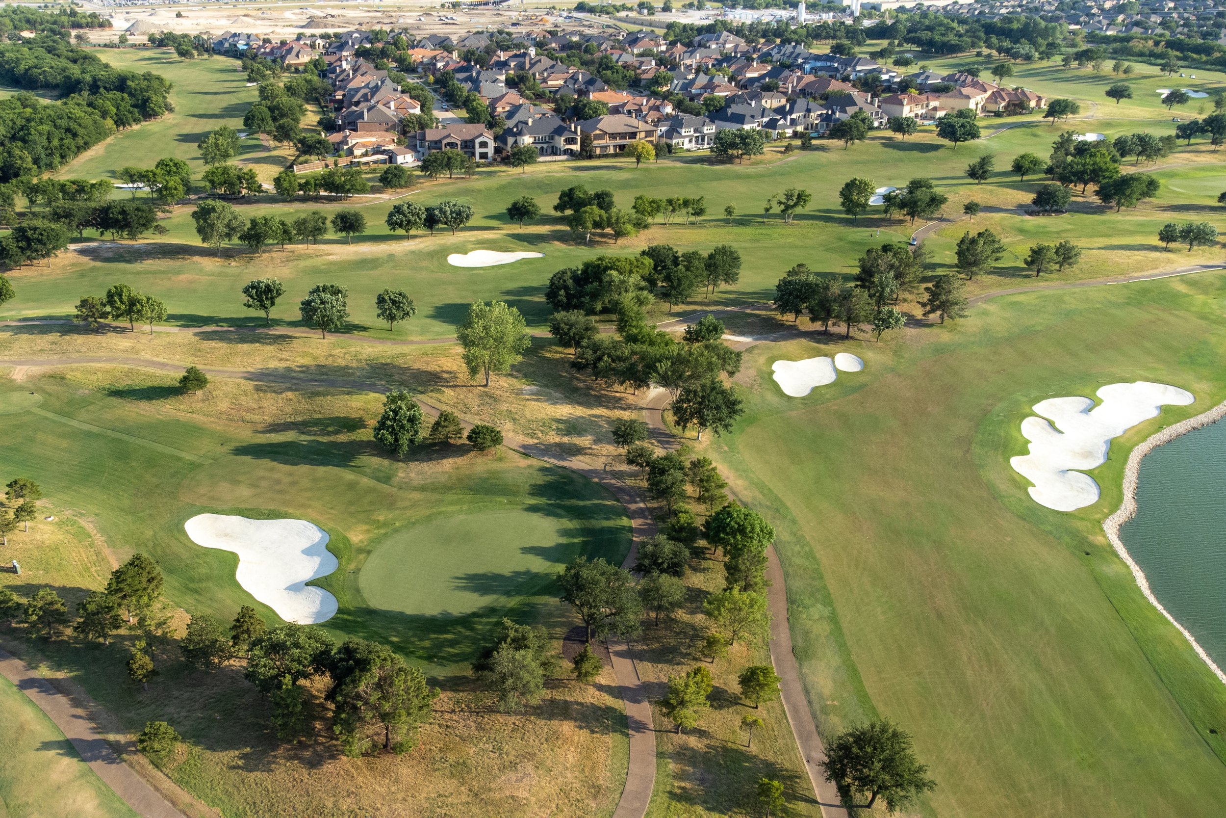 Aerial view of a golf course with multiple sand bunkers, green patches, trees, a water hazard on the right, paths, and a nearby residential neighborhood in the background.