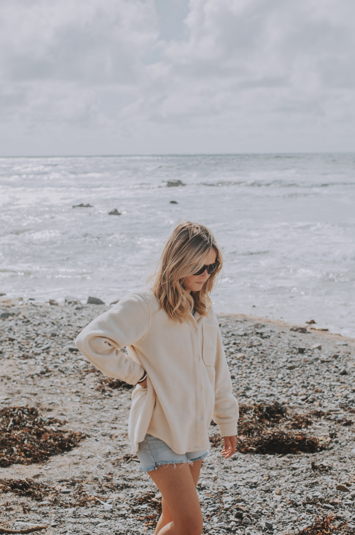 A woman with blonde hair wearing sunglasses, a cream-colored fleece jacket, and denim shorts standing on a rocky beach with ocean waves and cloudy sky in the background.
