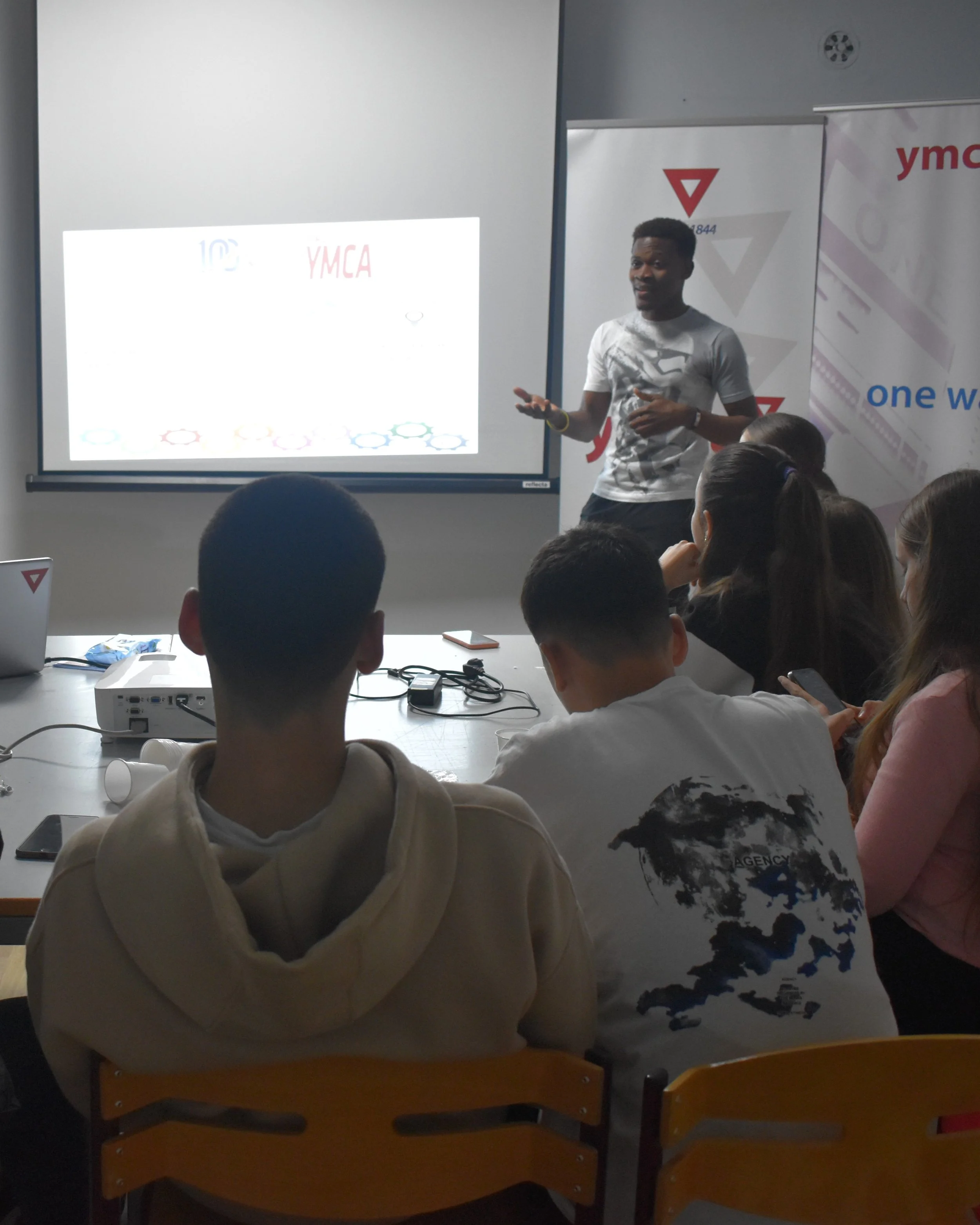 A man giving a presentation to a group of students in a classroom or seminar room with YMCA banners and a projector screen.