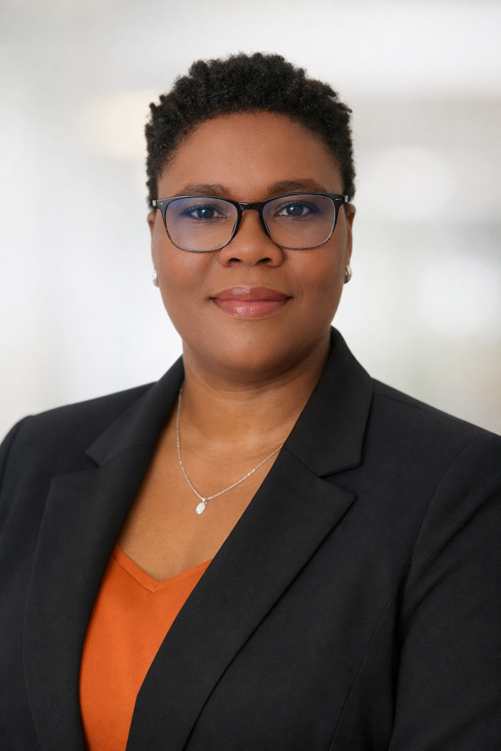 A professional woman with short curly hair, glasses, wearing a black blazer, orange blouse, and a necklace, posing against a plain background.