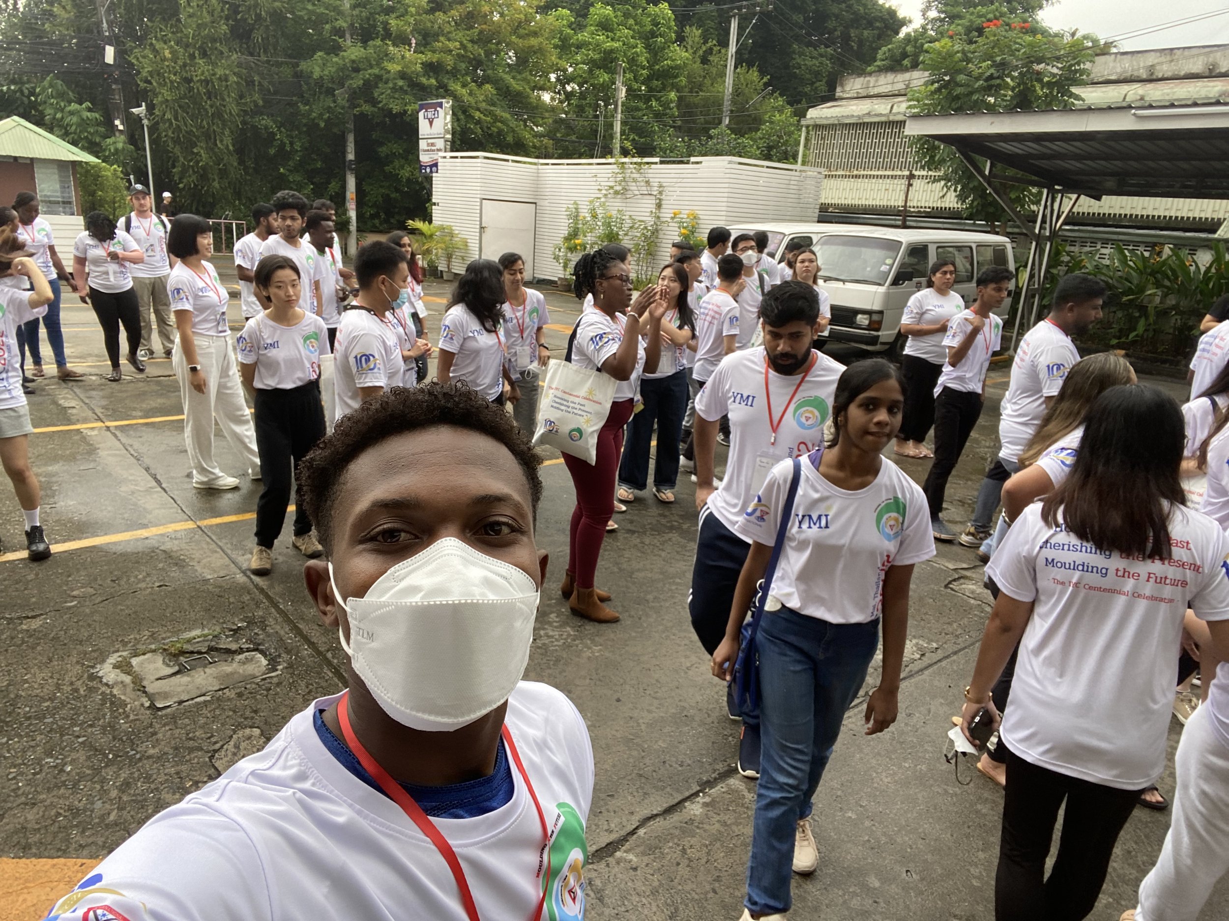 A group of diverse people, mostly wearing white t-shirts with logos, standing outdoors in a parking lot, participating in an event. Some are wearing face masks, and they are arranged in lines or small groups.