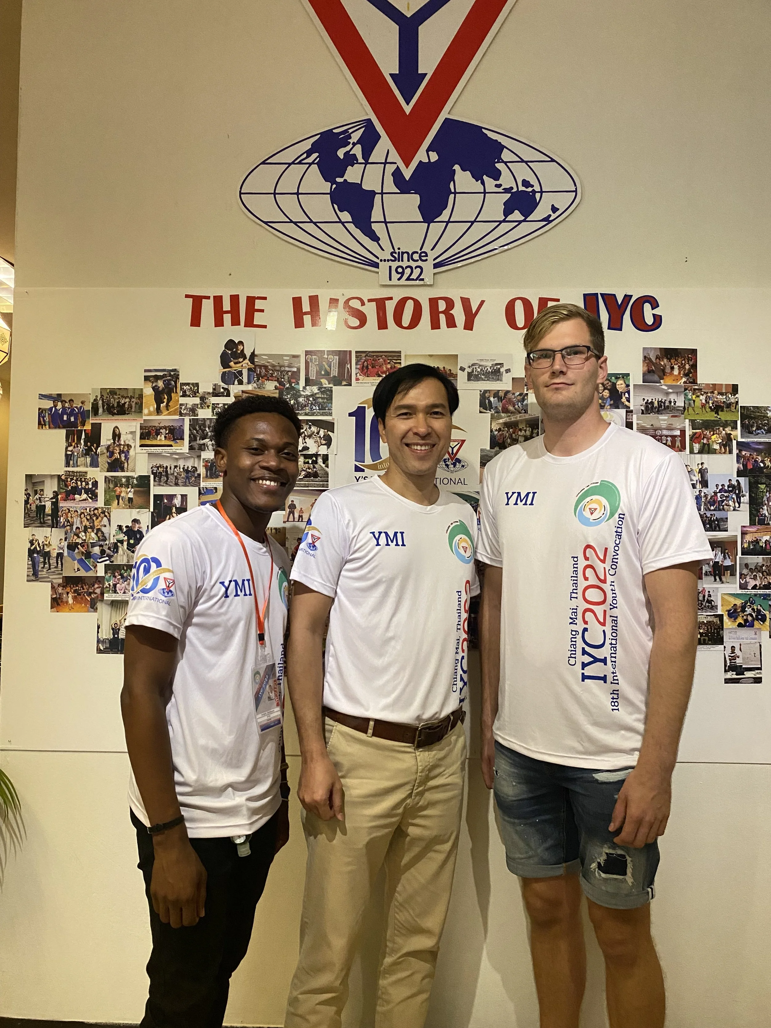Three men standing in front of a wall with photos, a banner that says 'The History of IYC,' and a large emblem with a globe and the text 'Since 1922.' They are wearing white T-shirts with the event details and logos.