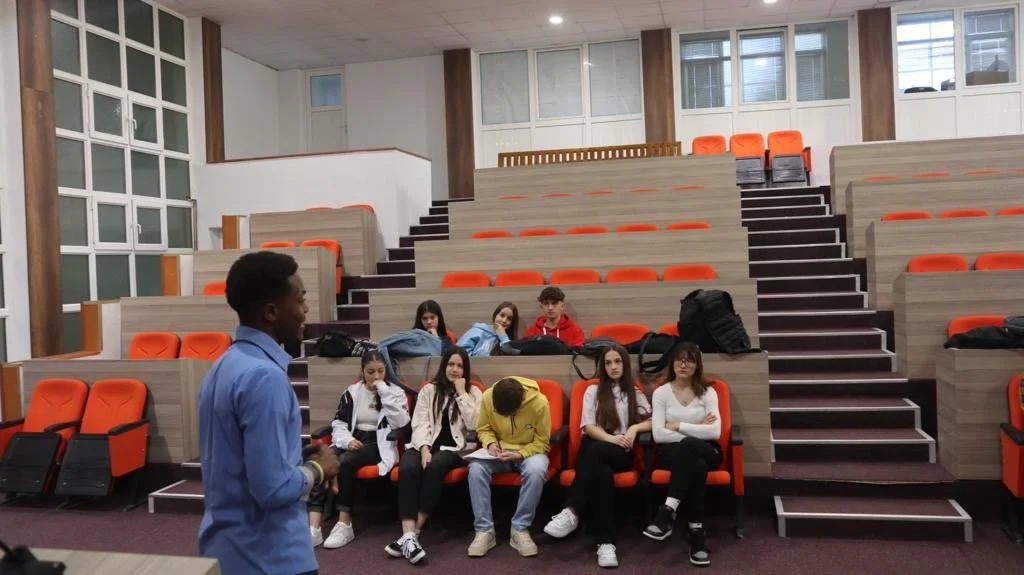A young man is speaking to a group of students seated in a tiered lecture hall with orange seats. The students are listening attentively, some taking notes and others looking at the speaker.