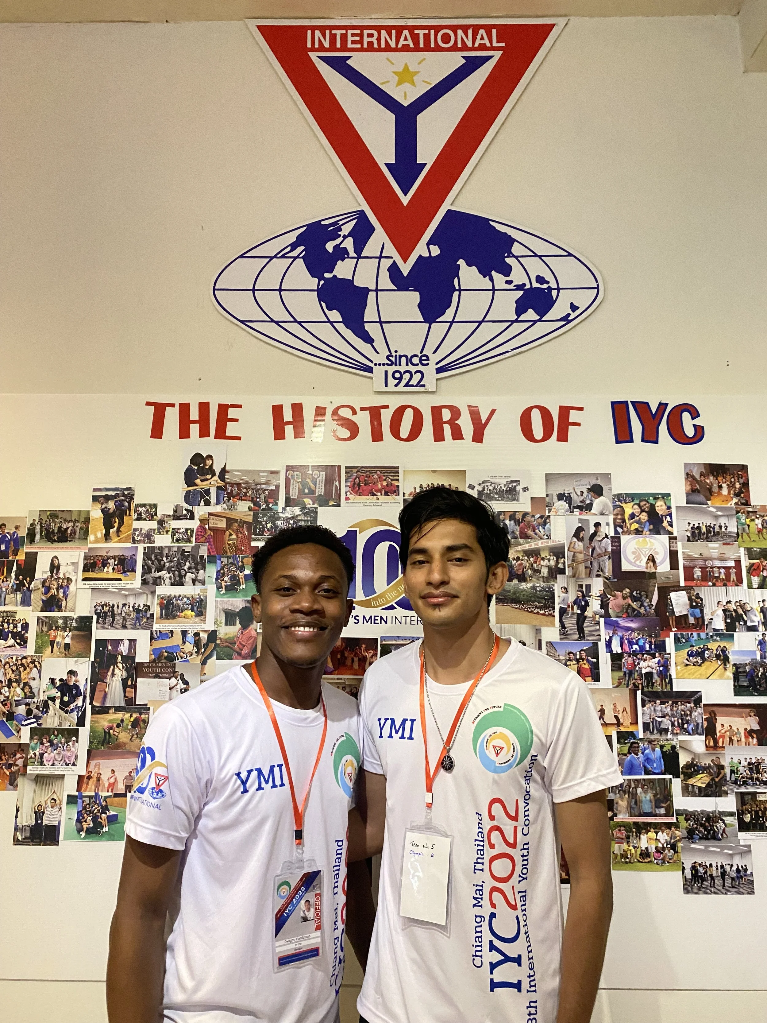 Two young men wearing white T-shirts and medals, standing in front of a collage of photos and a banner with the logo and branding of the International Youth Congress in Chiang Mai, Thailand, 2022.