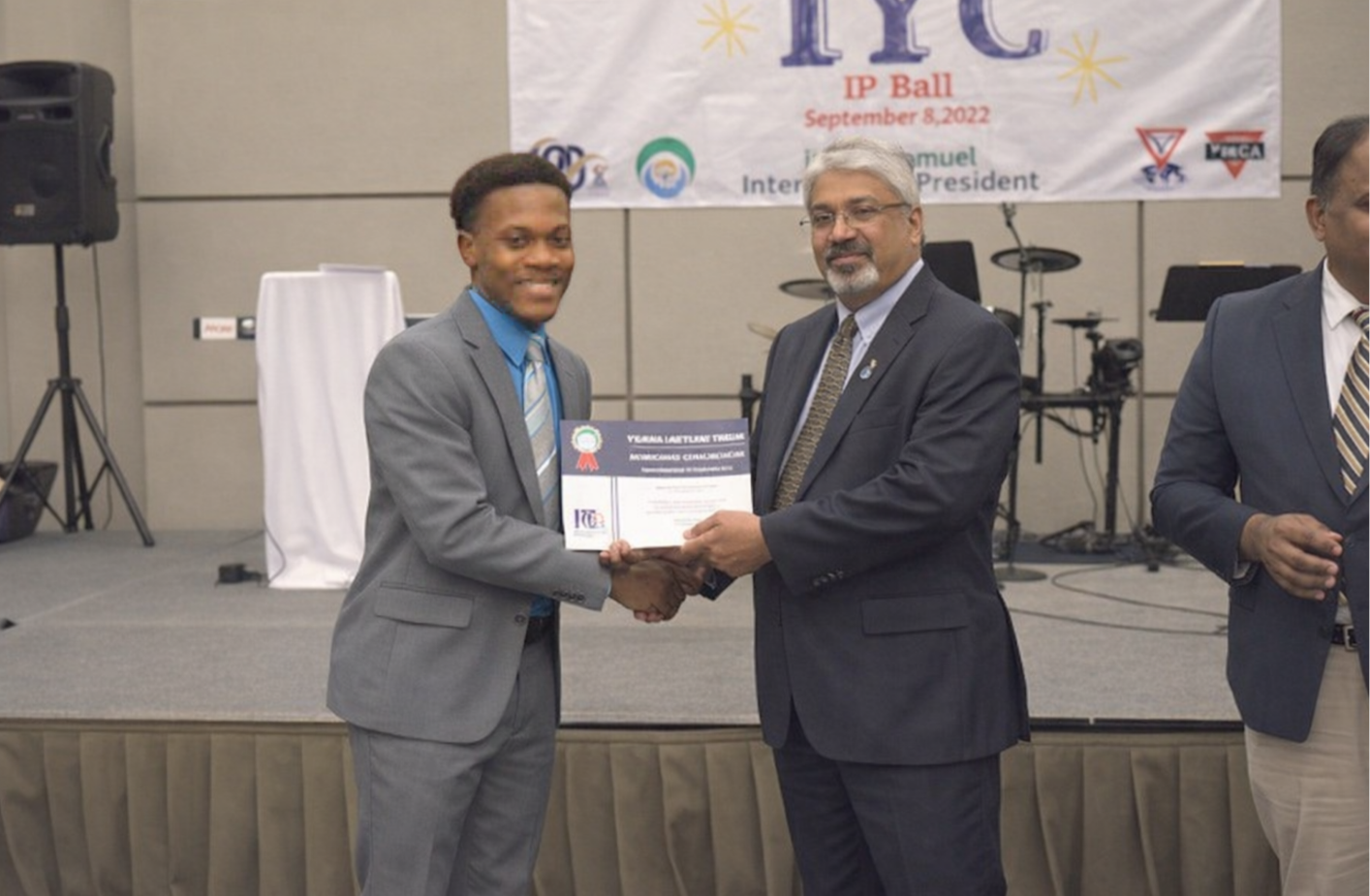 Two men in suits shaking hands during an award ceremony, with a certificate being handed over; a banner in the background indicates an event on September 8, 2022.