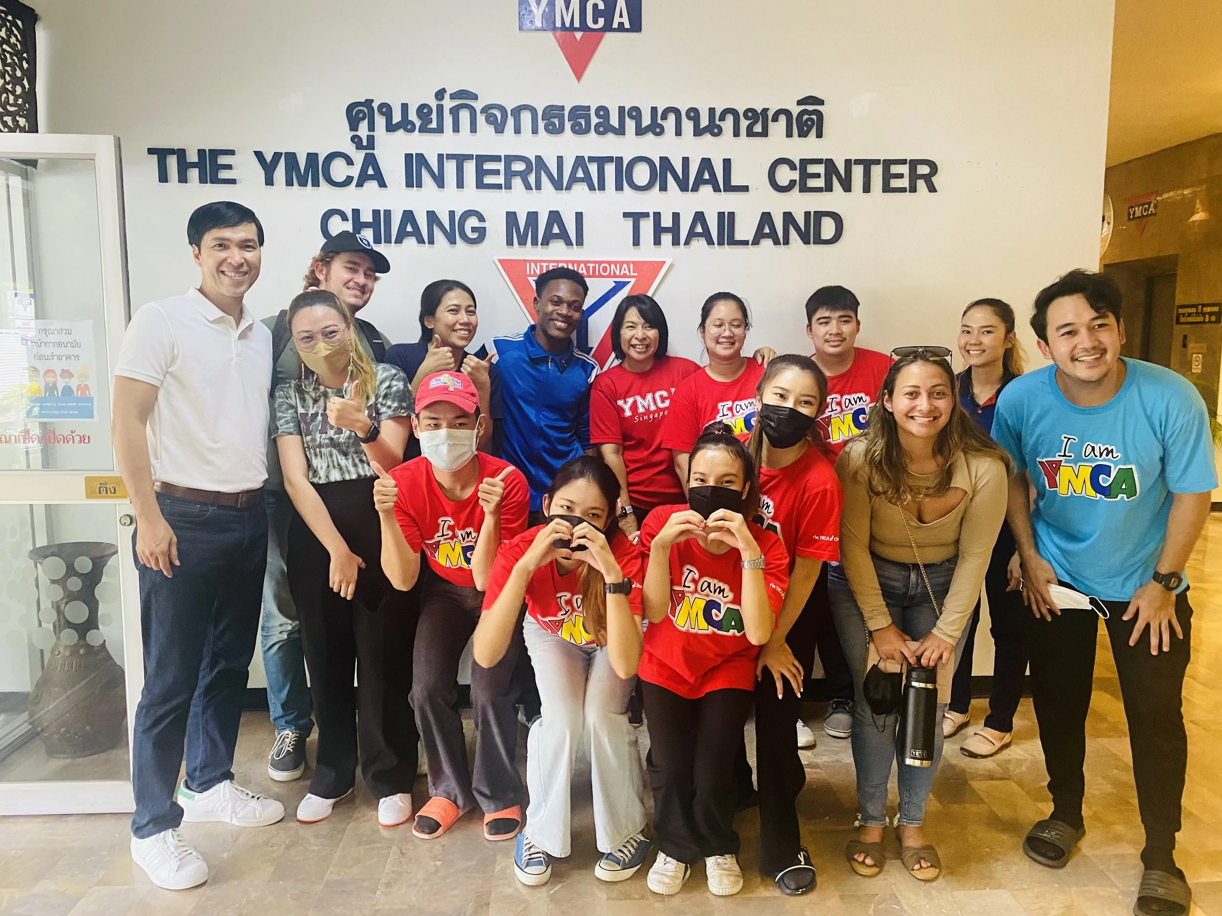 Group of people standing in front of a wall with the sign 'The YMCA International Center Chiang Mai Thailand', some wearing red shirts and others wearing blue and white shirts, smiling and posing for a photo.