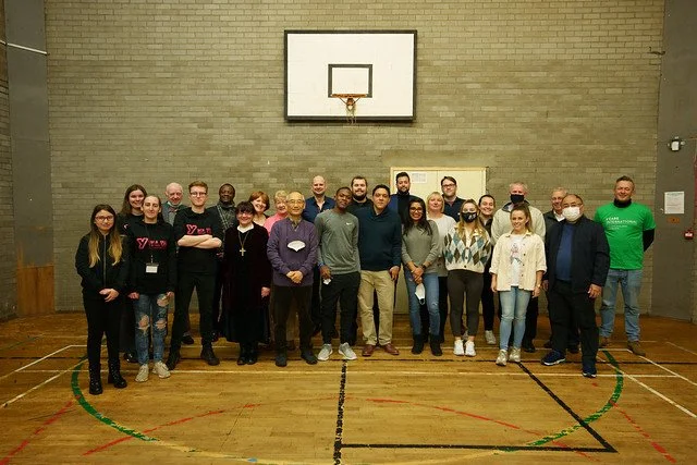 Group of 20 people standing in a gymnasium with a basketball hoop on the wall behind them.