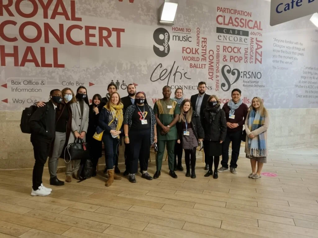 Group of people standing in front of a large wall with event information at the Royal Concert Hall, Glasgow. The background includes words related to music, dance, and cultural activities.