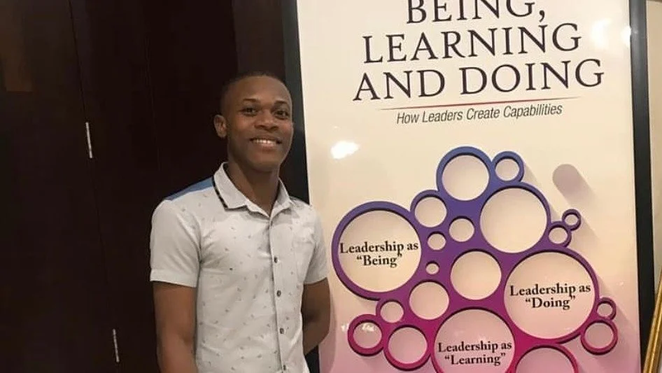 A smiling young man standing next to a large poster with the title "Being, Learning and Doing" and the subtitle "How Leaders Create Capabilities." The poster features circles with the words "Leadership as 'Being', Leadership as 'Doing', Leadership as