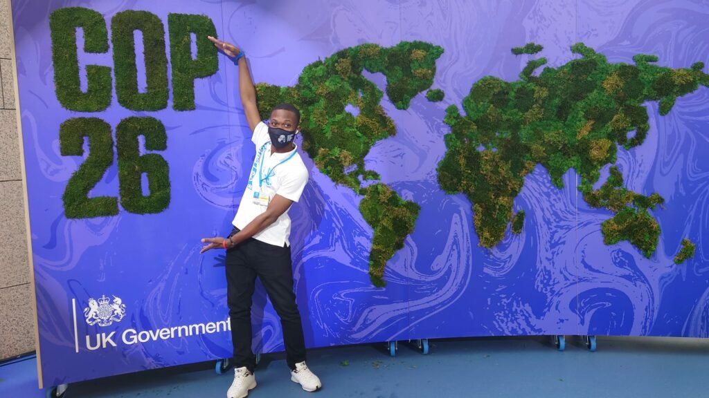 A person wearing a face mask and white T-shirt standing in front of a large blue backdrop with a world map made of green foliage. The backdrop features the text 'COP 26' and the UK Government logo.