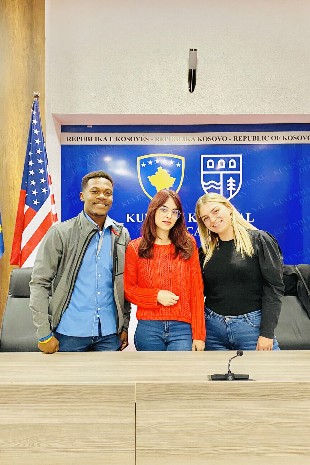 Three young women standing together in front of a Kosovo government backdrop inside a conference room, with an American flag to the left.