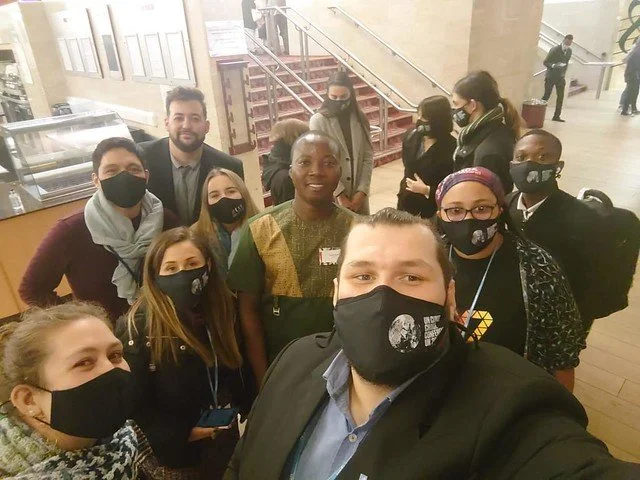 Group of people wearing masks, taking a selfie in an indoor setting with a staircase in the background.