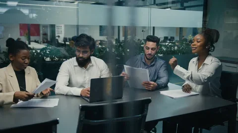 Four diverse professionals sitting at a conference table in a modern office, engaged in a discussion with documents and a laptop.