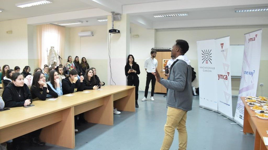 A young man giving a presentation to a group of students seated at long tables in a classroom or conference room with banners in the background.