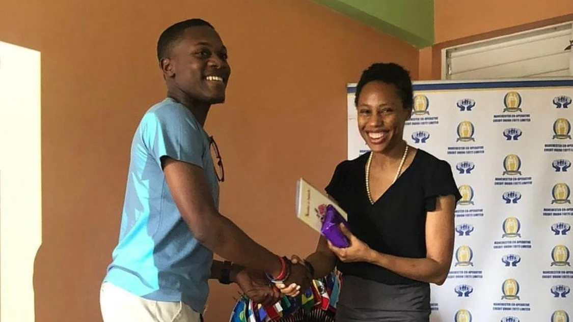 A man and a woman shaking hands and smiling at each other, with the woman holding a gift bag and a book, in front of a banner with logos.