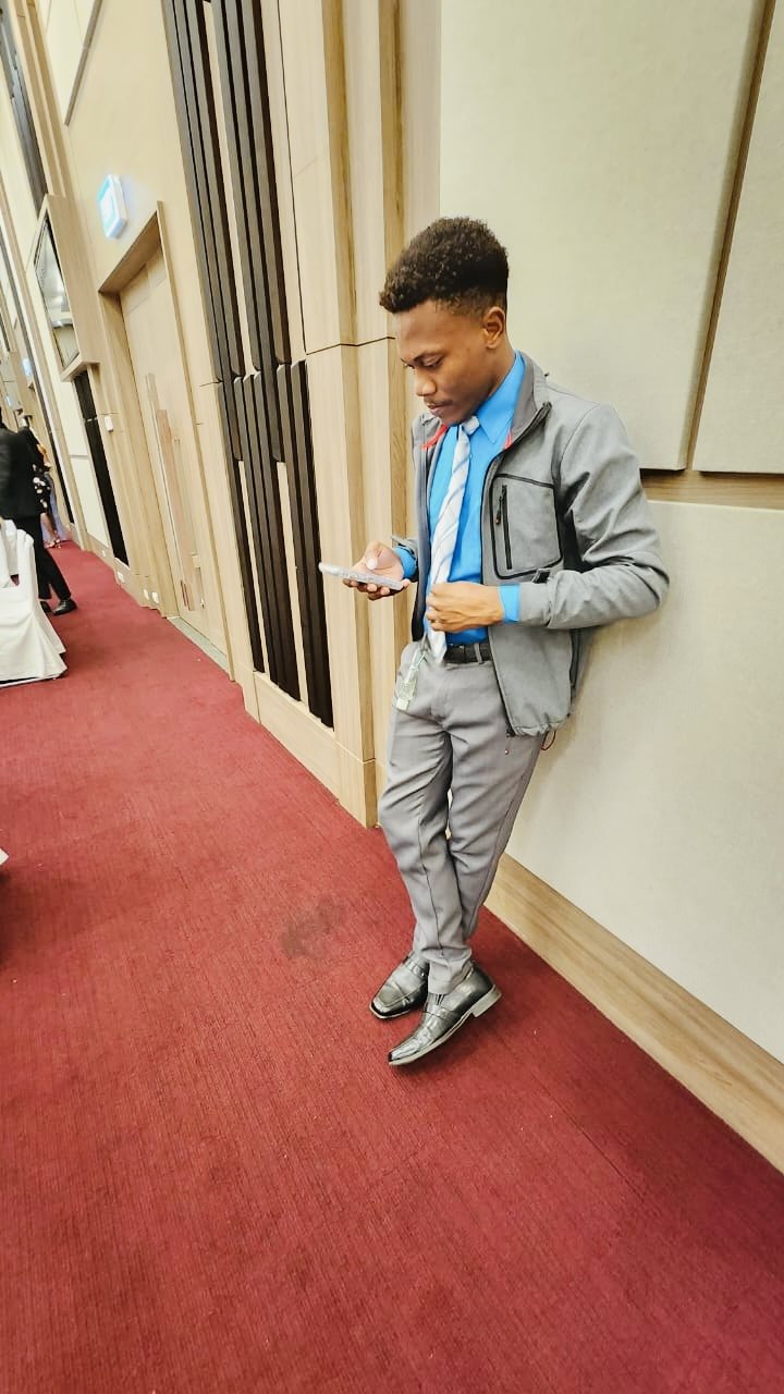 Young man in gray suit, blue shirt, and tie standing against wall, looking at his phone in a conference or event hall.