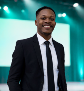 A smiling man in a suit standing in front of a lit stage or screen.
