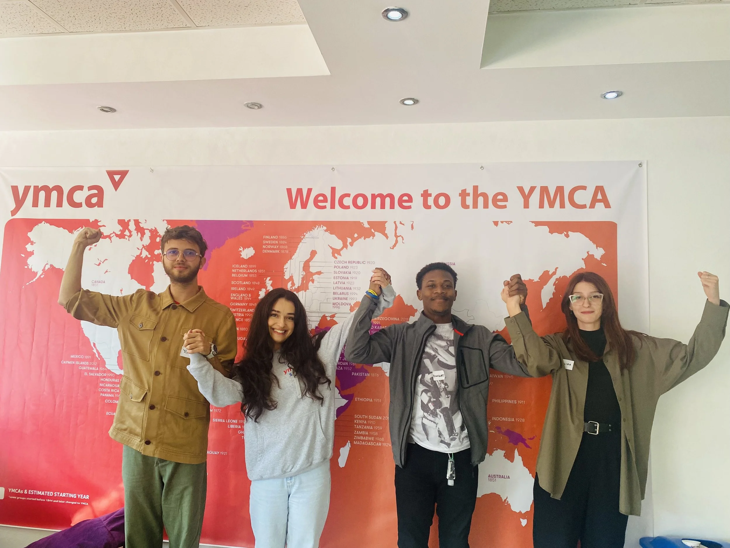 Four young adults, two men and two women, standing in front of a large world map with a YMCA banner, raising their arms in a celebratory pose.