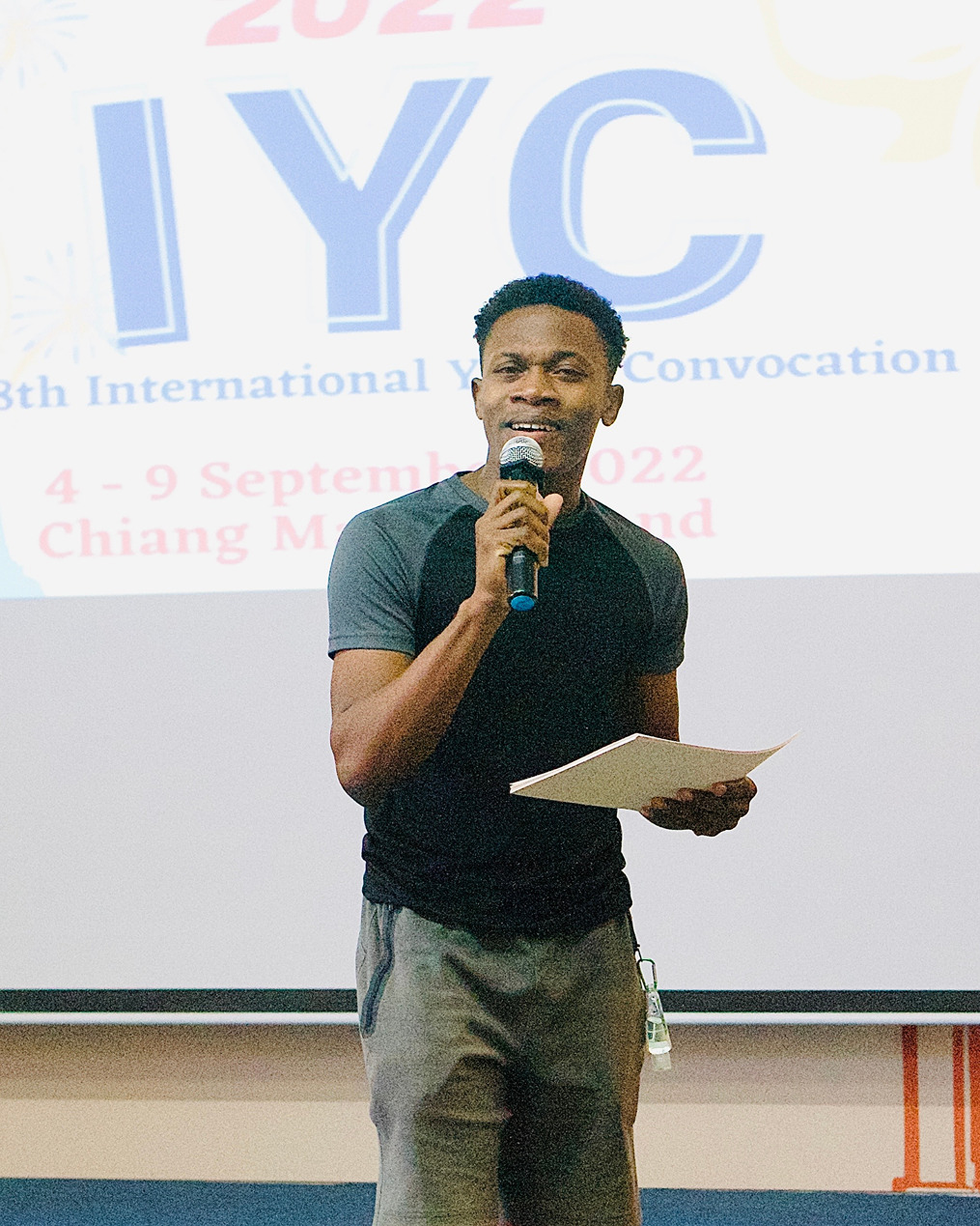 A man speaking into a microphone while holding a paper, standing in front of a screen that displays the logo for the 38th International Youth Convocation, held from September 4-9, 2022, in Chiang Mai, Thailand.