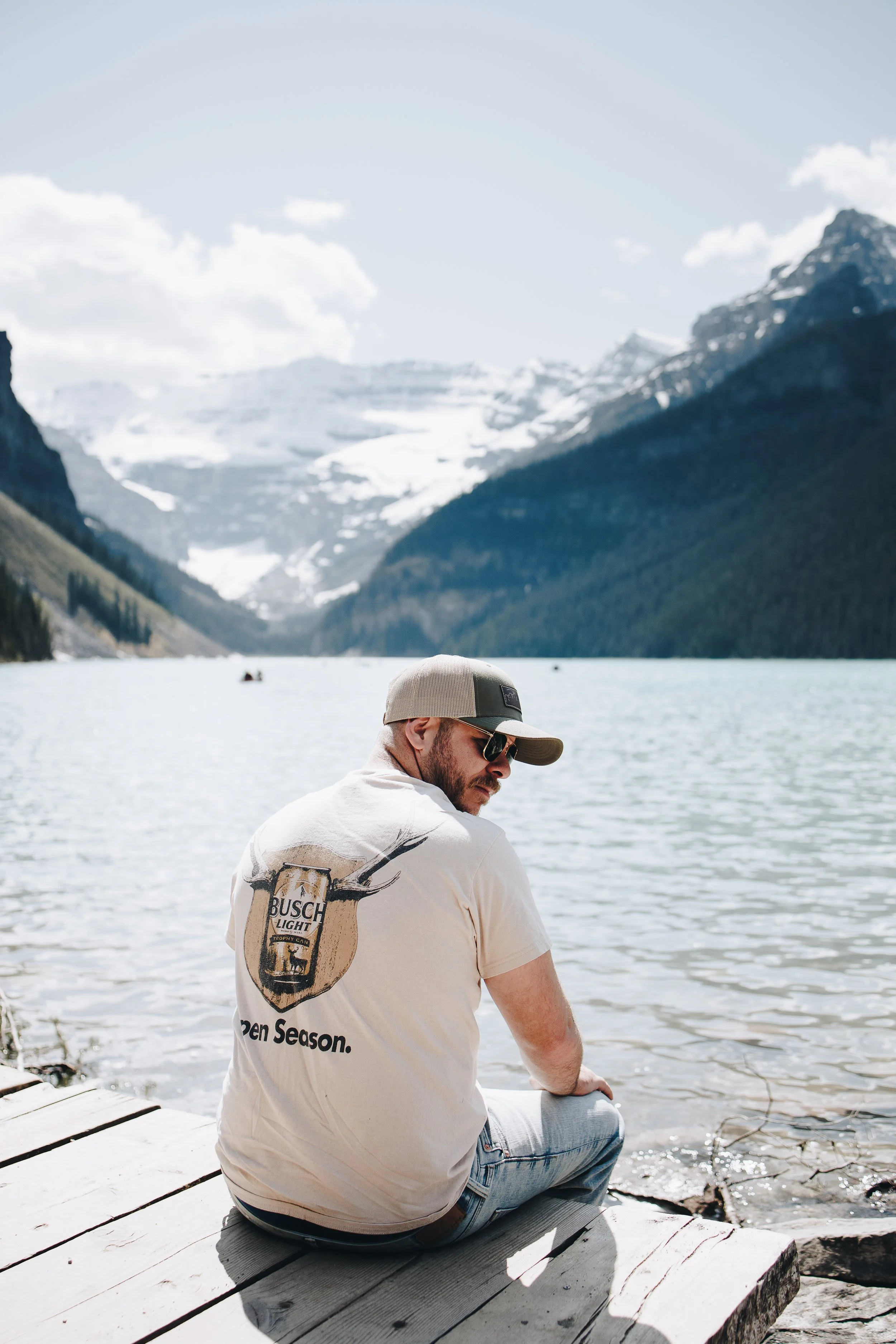 A man sitting on a wooden dock beside a lake with mountains and snow in the background, wearing a beige t-shirt with a Busch Light logo and a baseball cap.