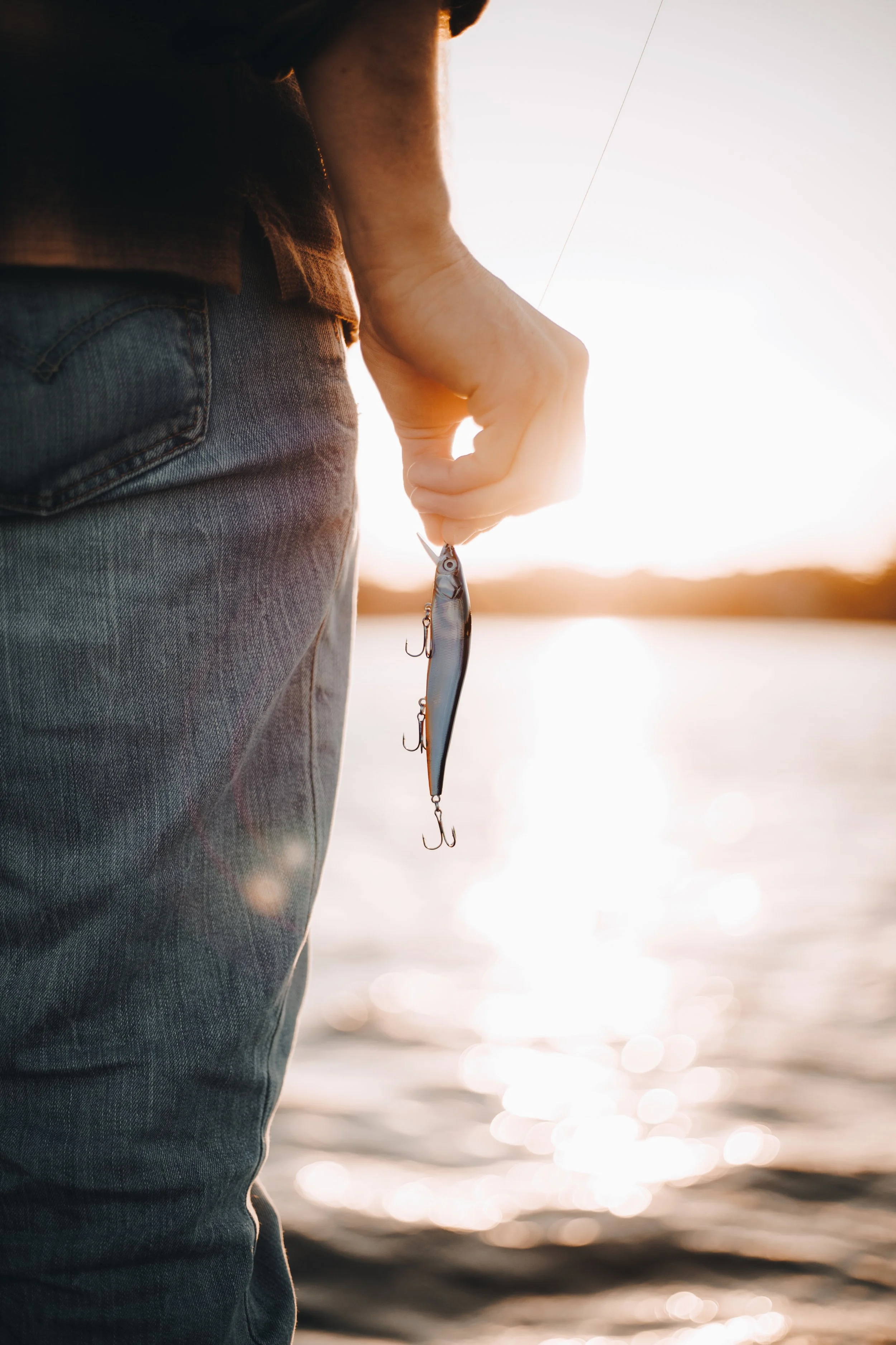 Close-up of a person holding a fishing lure by the water at sunset.