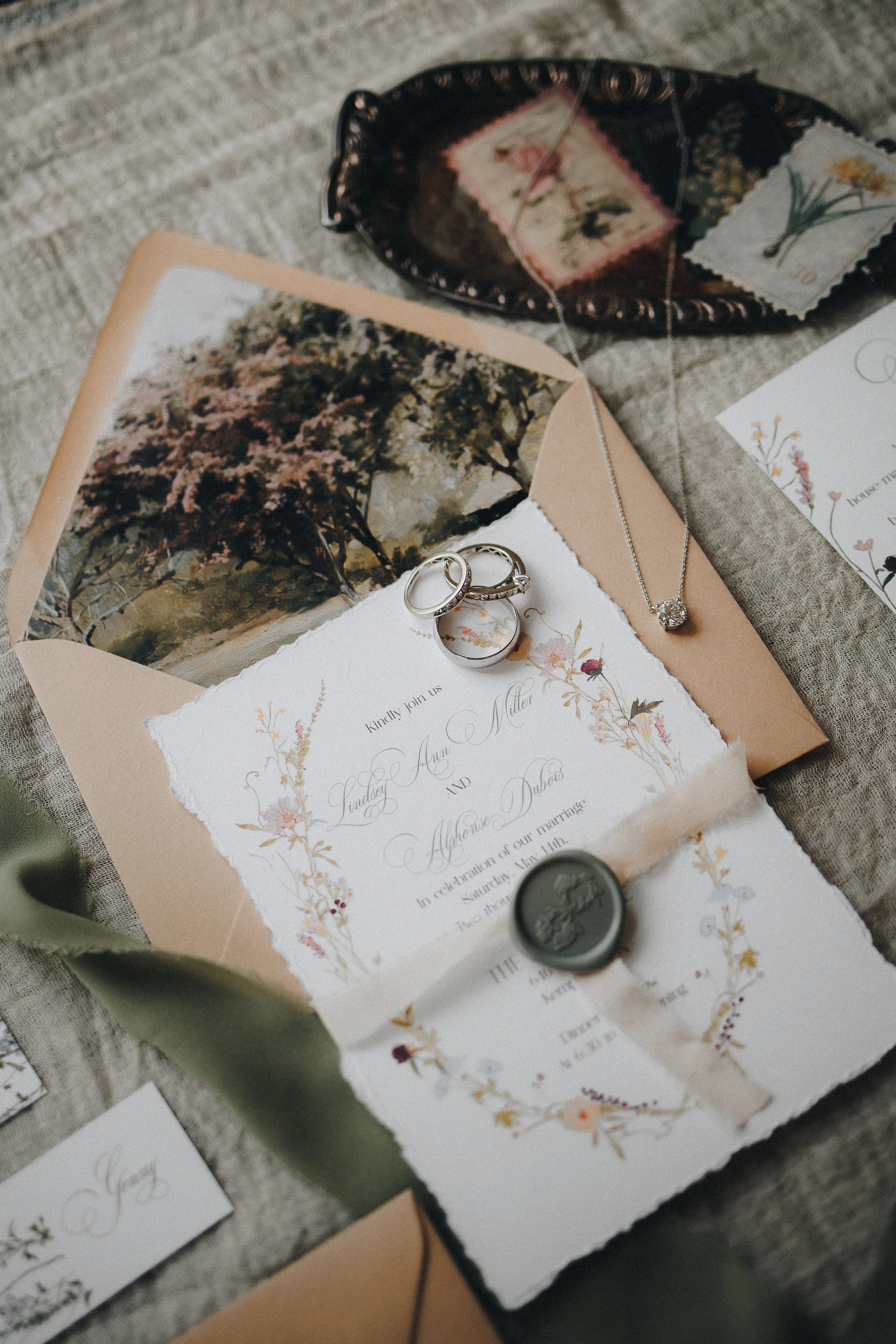Wedding invitation with floral design, wedding rings, and jewelry on textured fabric.