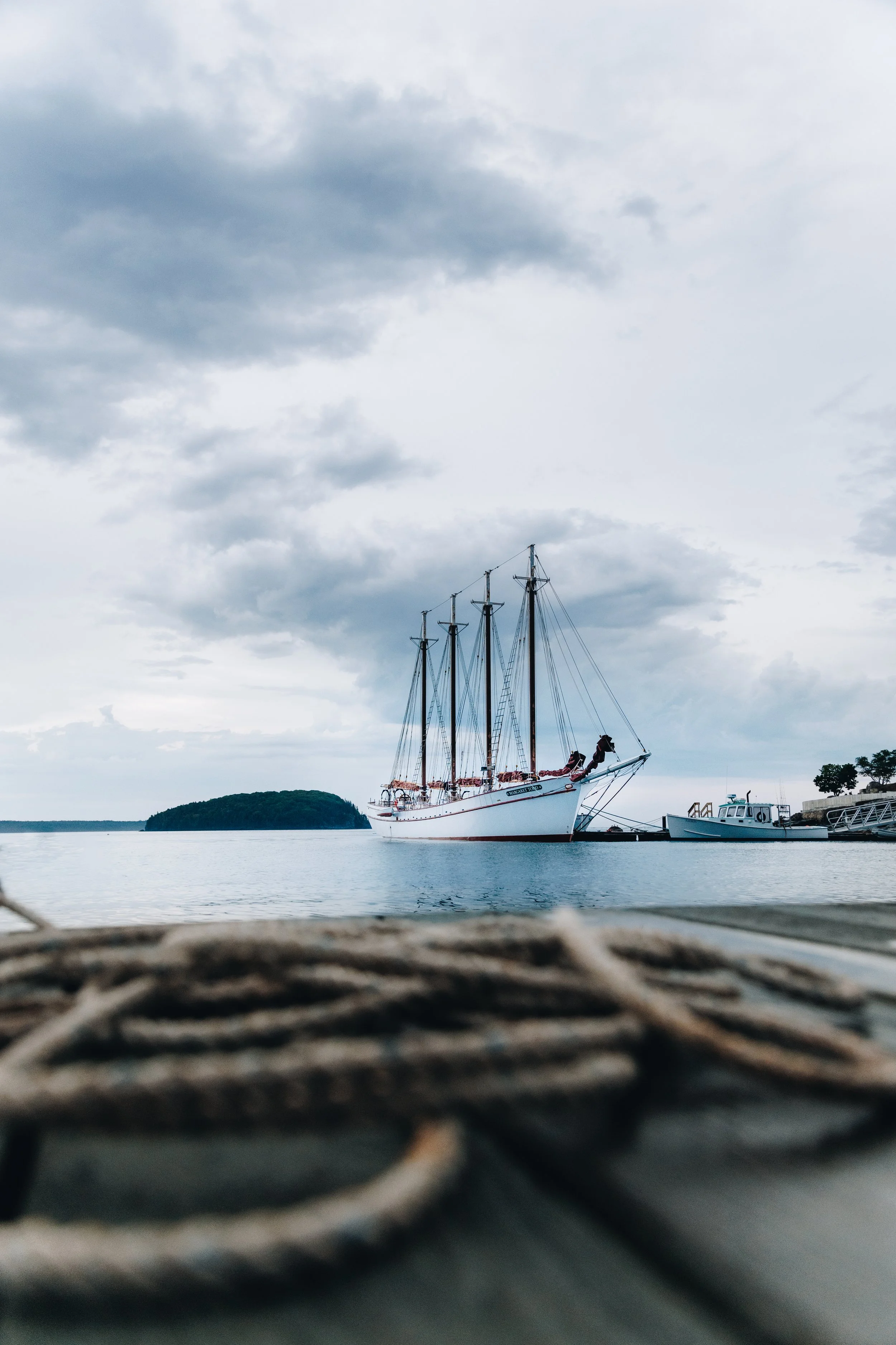 A large white sailing ship docked at a marina with smaller boats nearby, under a cloudy sky.