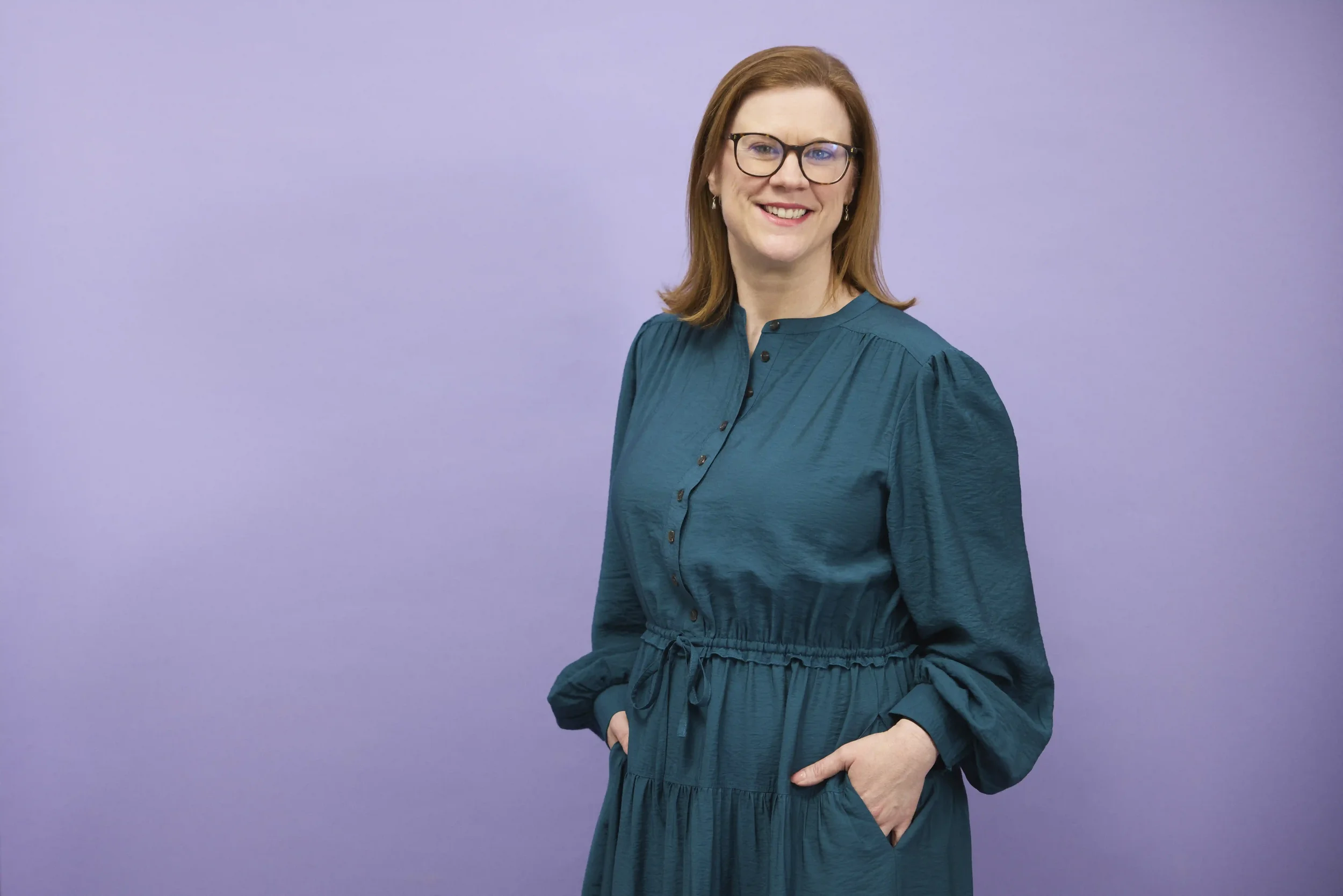 Woman with glasses smiling, wearing a teal dress with a cinched waist and long sleeves, standing against a plain light purple background.